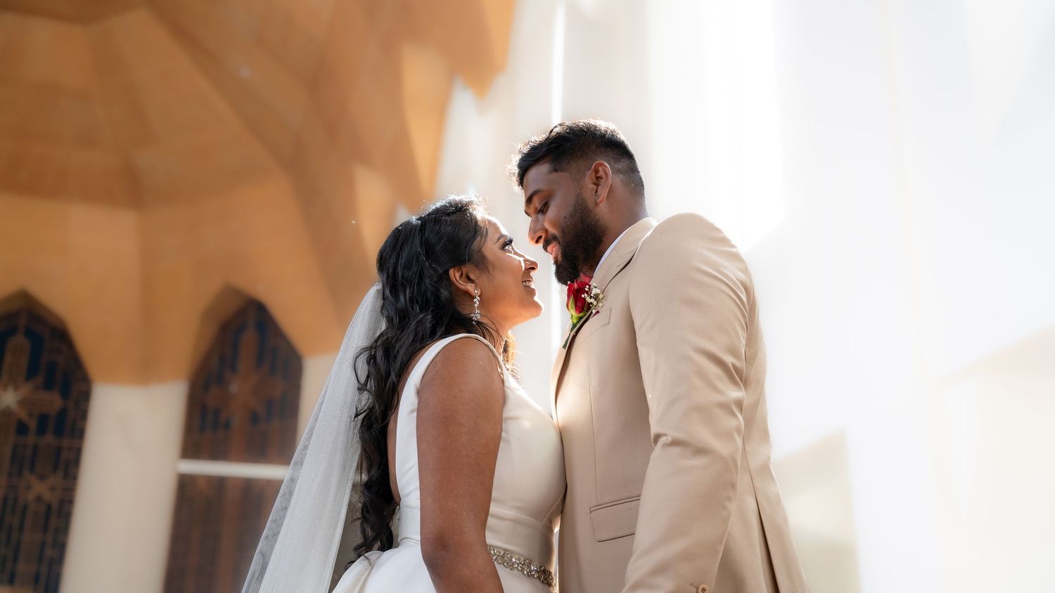 Couple in wedding attire share an intimate moment together inside a sunlit church with arched windows.