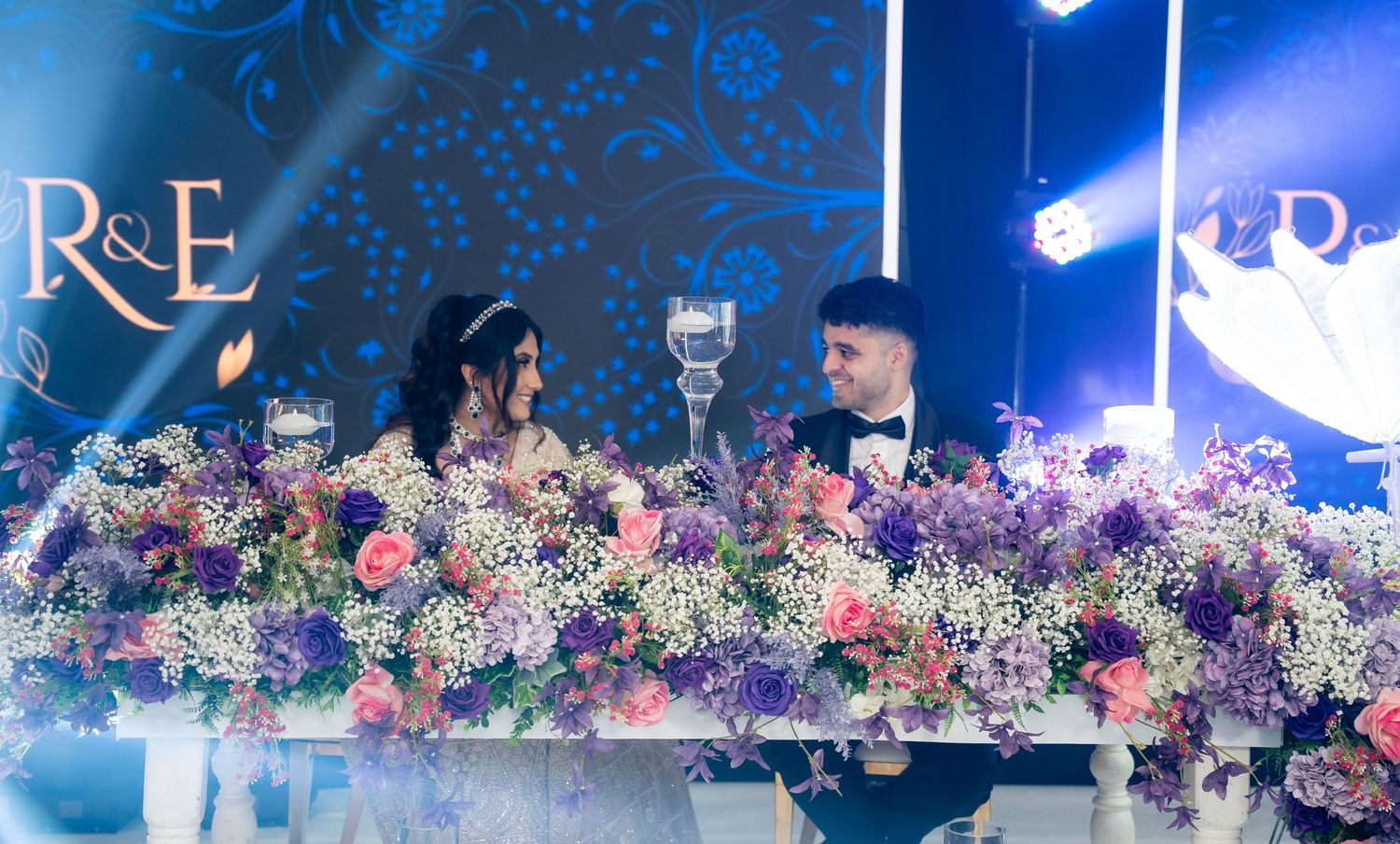 Bride and groom seated at floral wedding reception table with purple and pink flowers under blue decorative lighting.