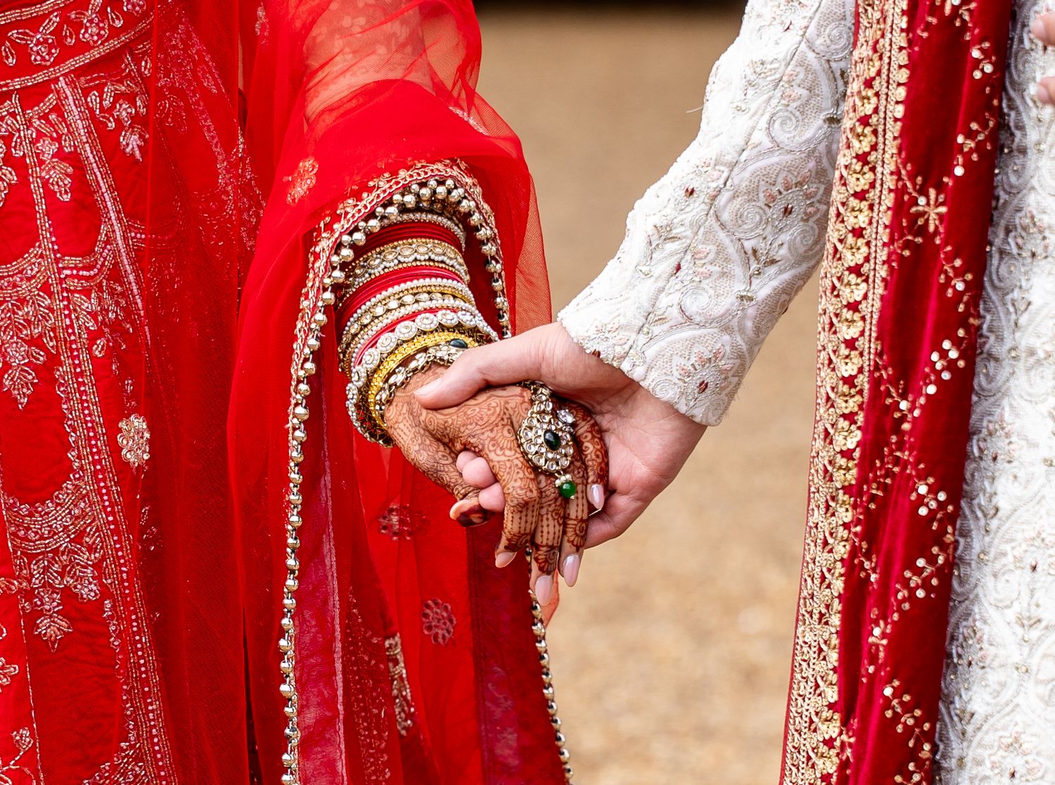 Close-up of bride and groom holding hands, bride wearing red lehenga with intricate mehndi and bangles.