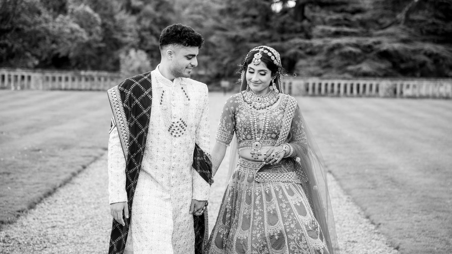Black and white photo of Indian bride and groom holding hands on a garden pathway.