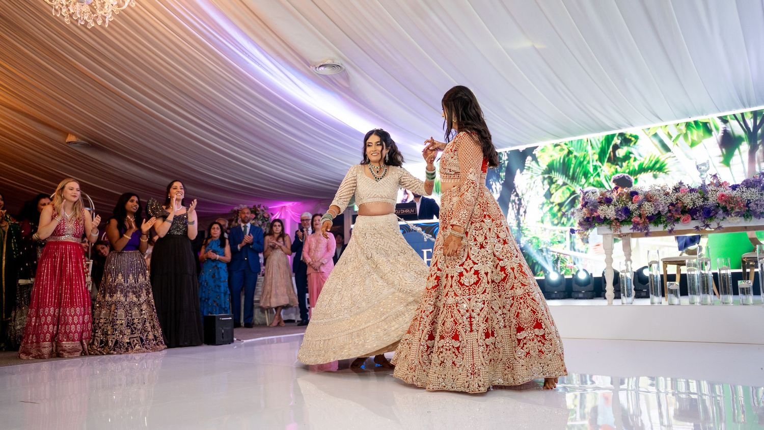 Two women in elegant Indian lehengas dancing at a wedding reception in a decorated banquet hall.