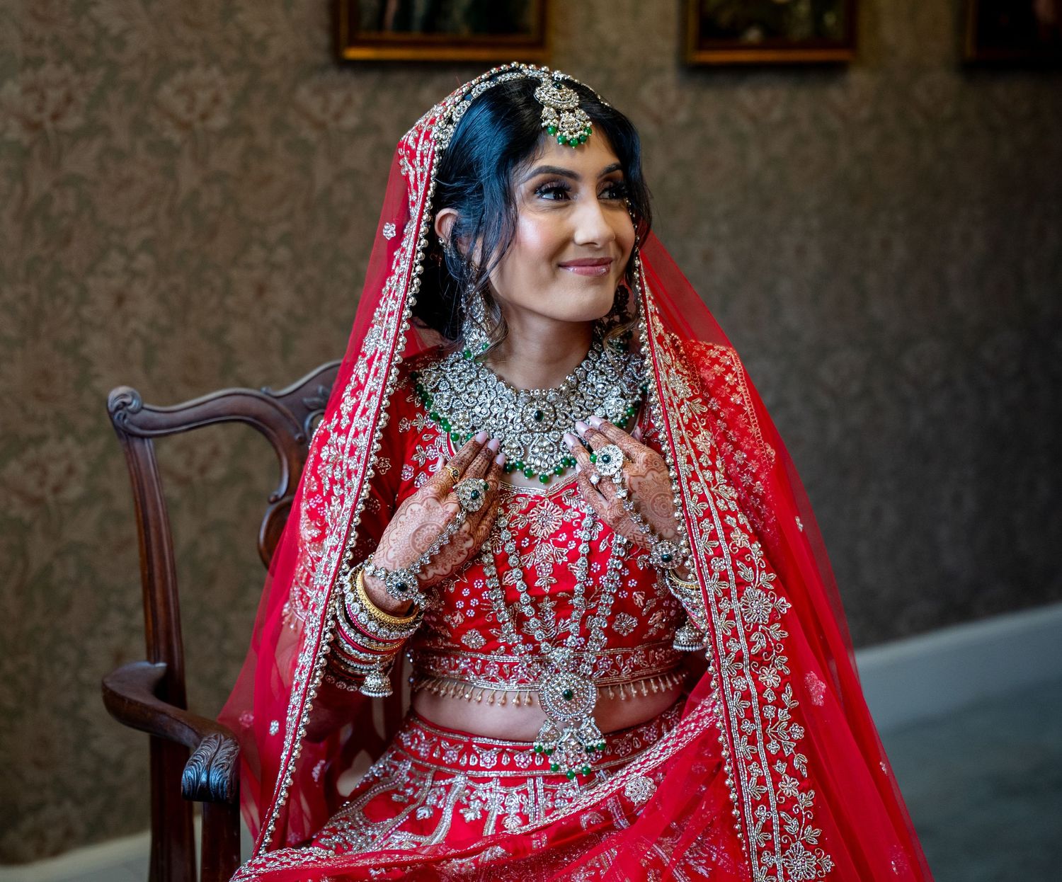 Indian bride in red bridal lehenga with heavy silver jewelry and dupatta seated on chair.