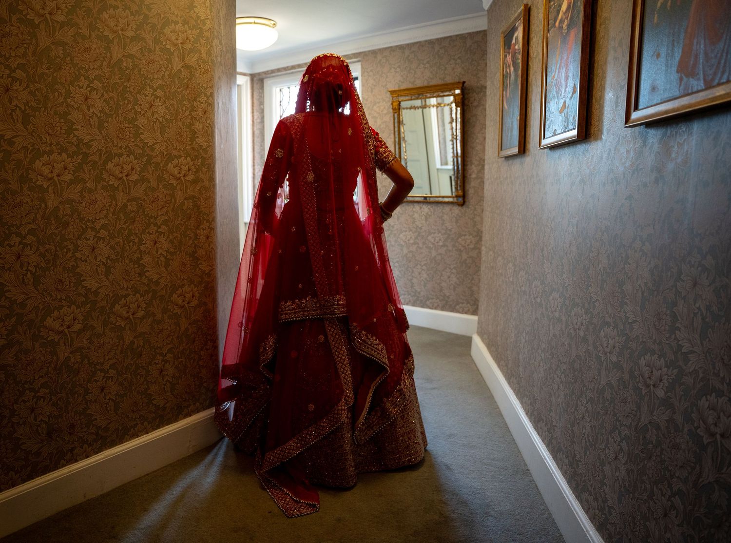 A bride in a stunning red lehenga and dupatta stands in a hallway, showcasing traditional South Asian bridal attire.