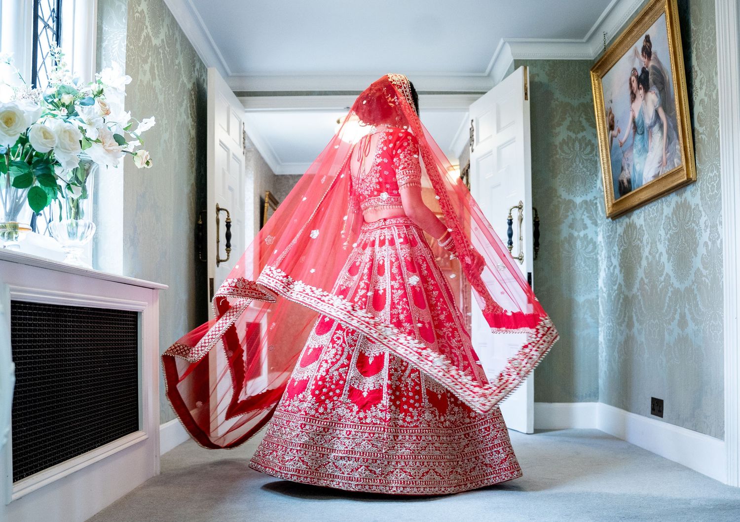 Indian bride in red lehenga spinning with dupatta flowing in elegant hallway.