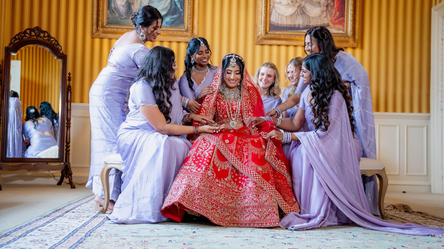 Bride in red lehenga surrounded by bridesmaids in lavender dresses during wedding preparation in elegant room.