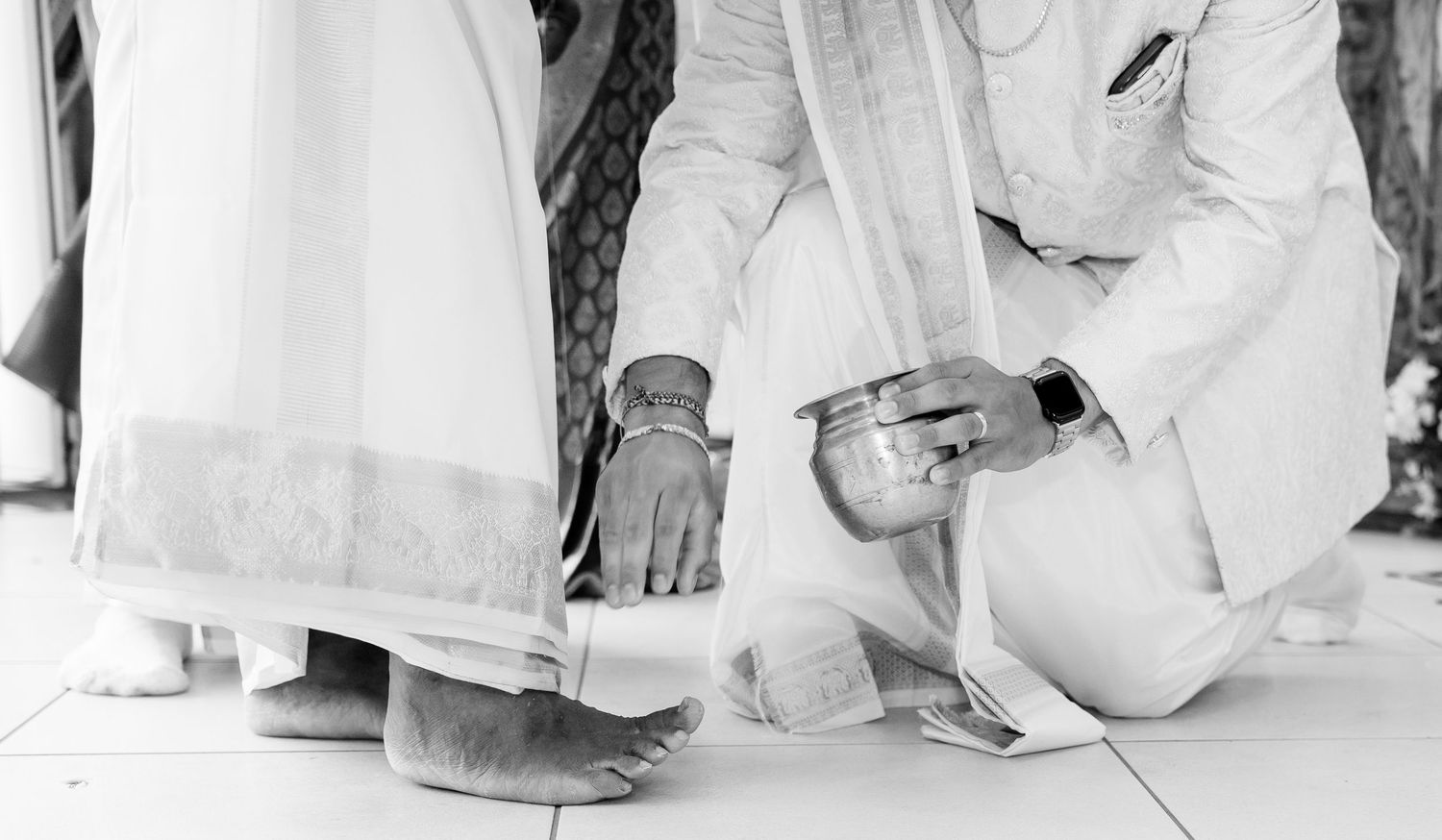 Groom kneeling to perform a traditional Hindu wedding ritual, washing the bride's feet with water from a ceremonial bowl.