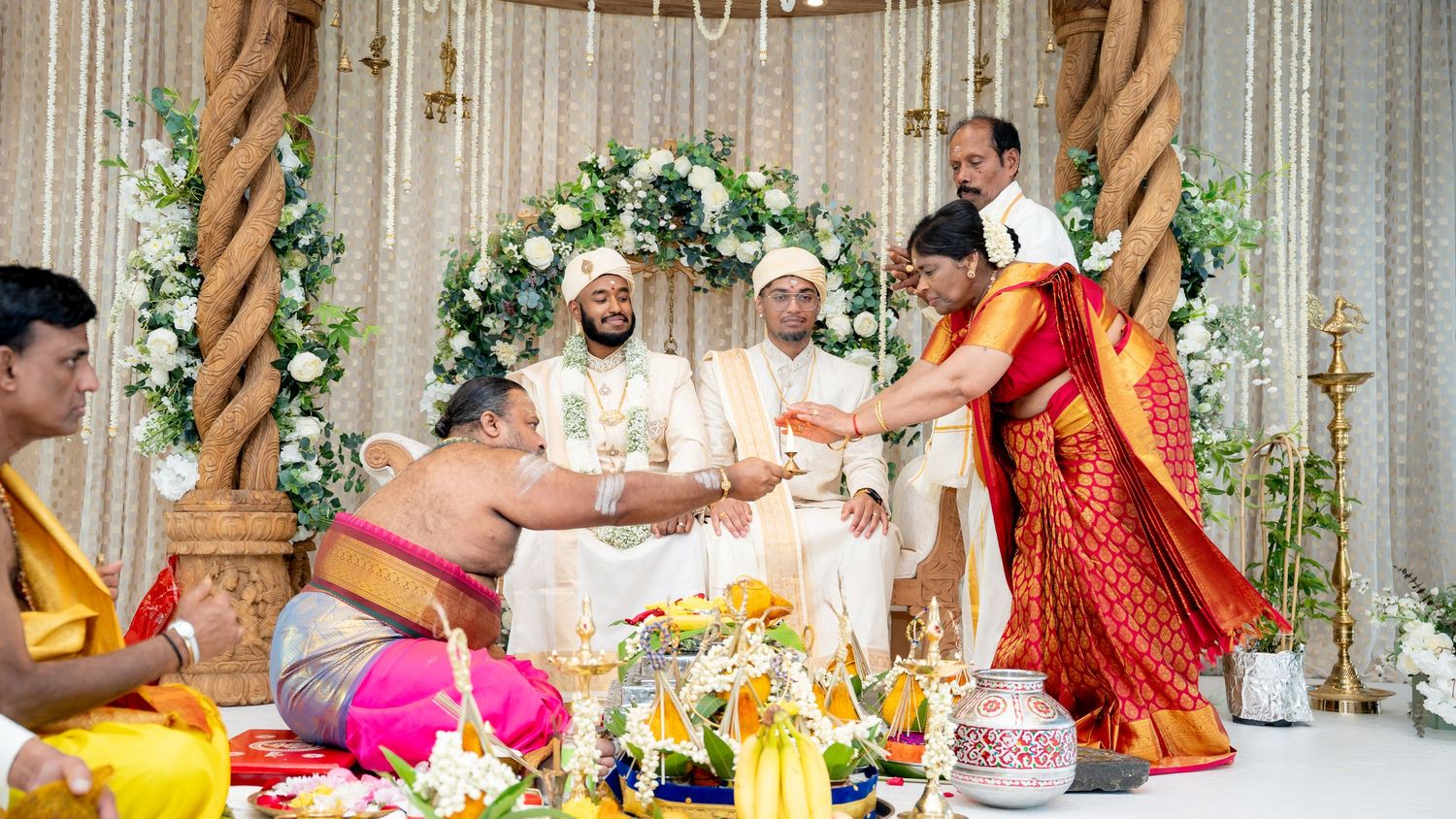 Traditional South Indian wedding ceremony with groom in white sherwani receiving blessings from priest at decorated mandap.
