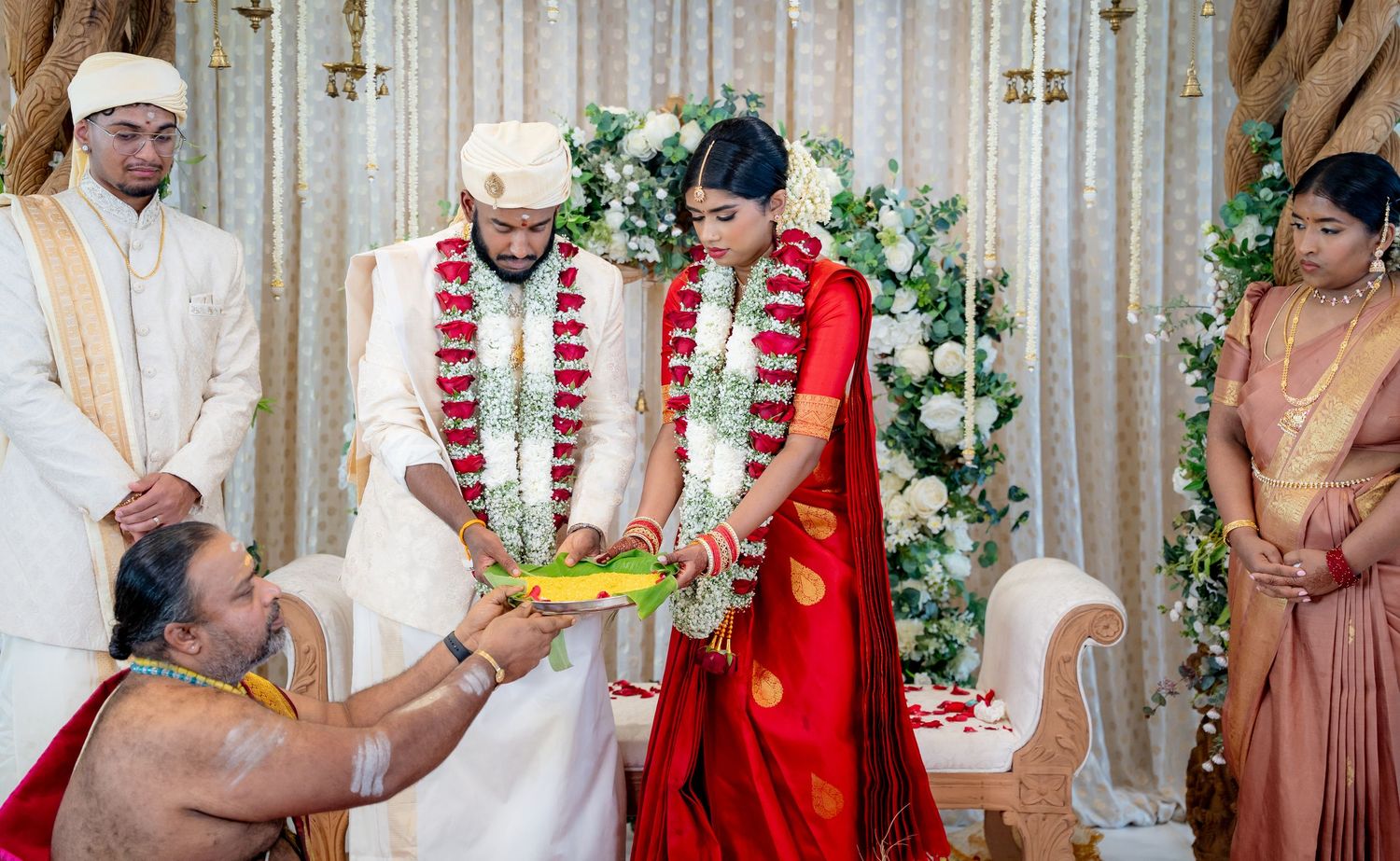 Hindu wedding ritual with priest presenting offerings to bride in red saree and groom in white dhoti.