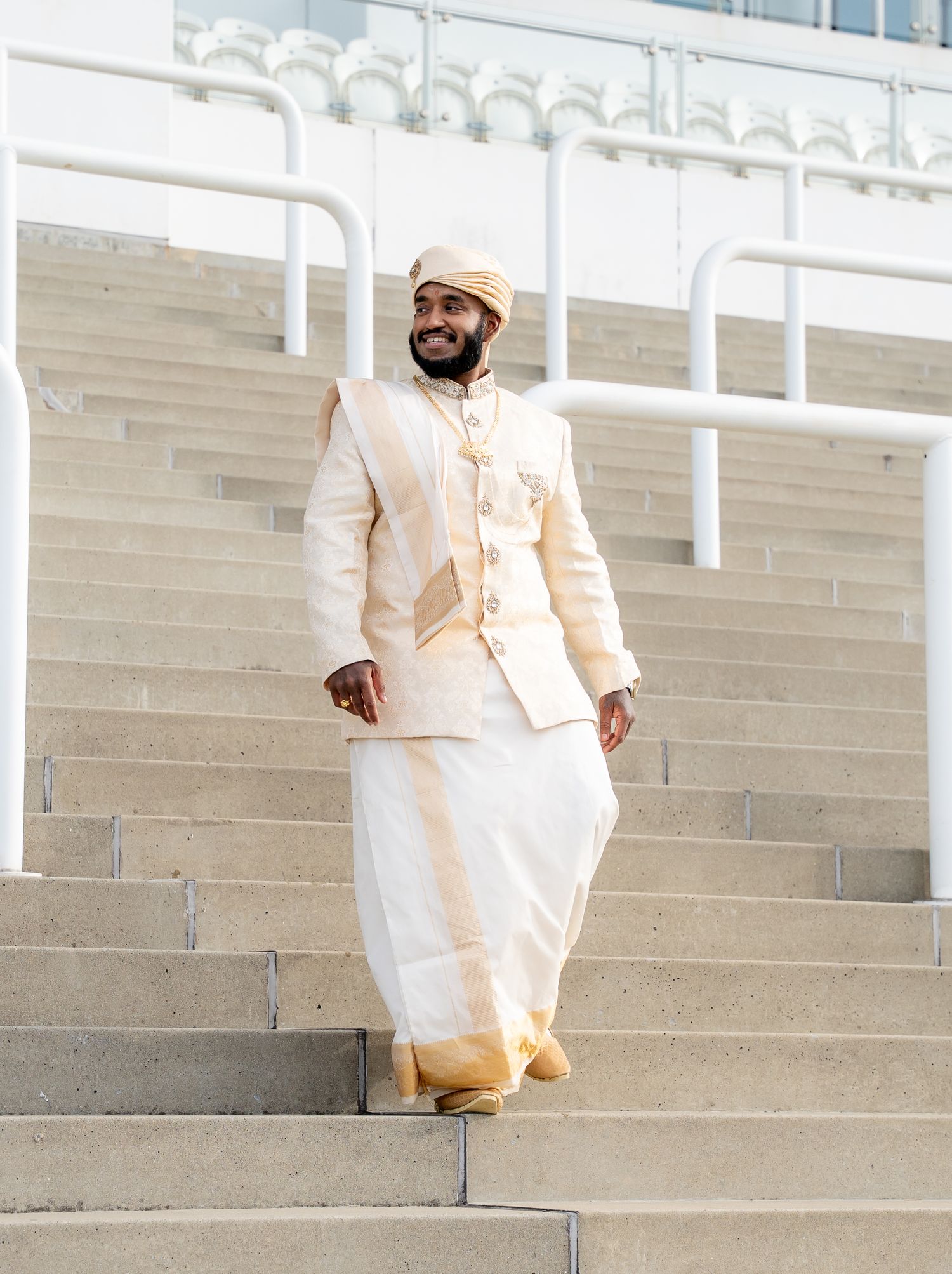 Smiling man in cream and gold traditional South Asian wedding outfit on stadium stairs.