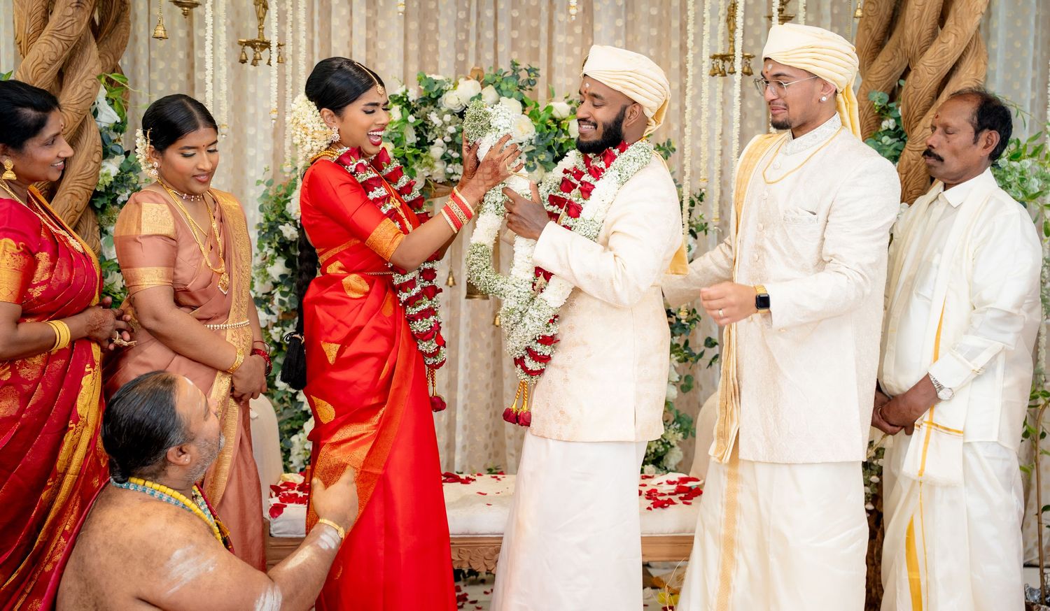 Traditional Hindu wedding garland exchange with bride in red Kanjivaram saree and groom in white attire.