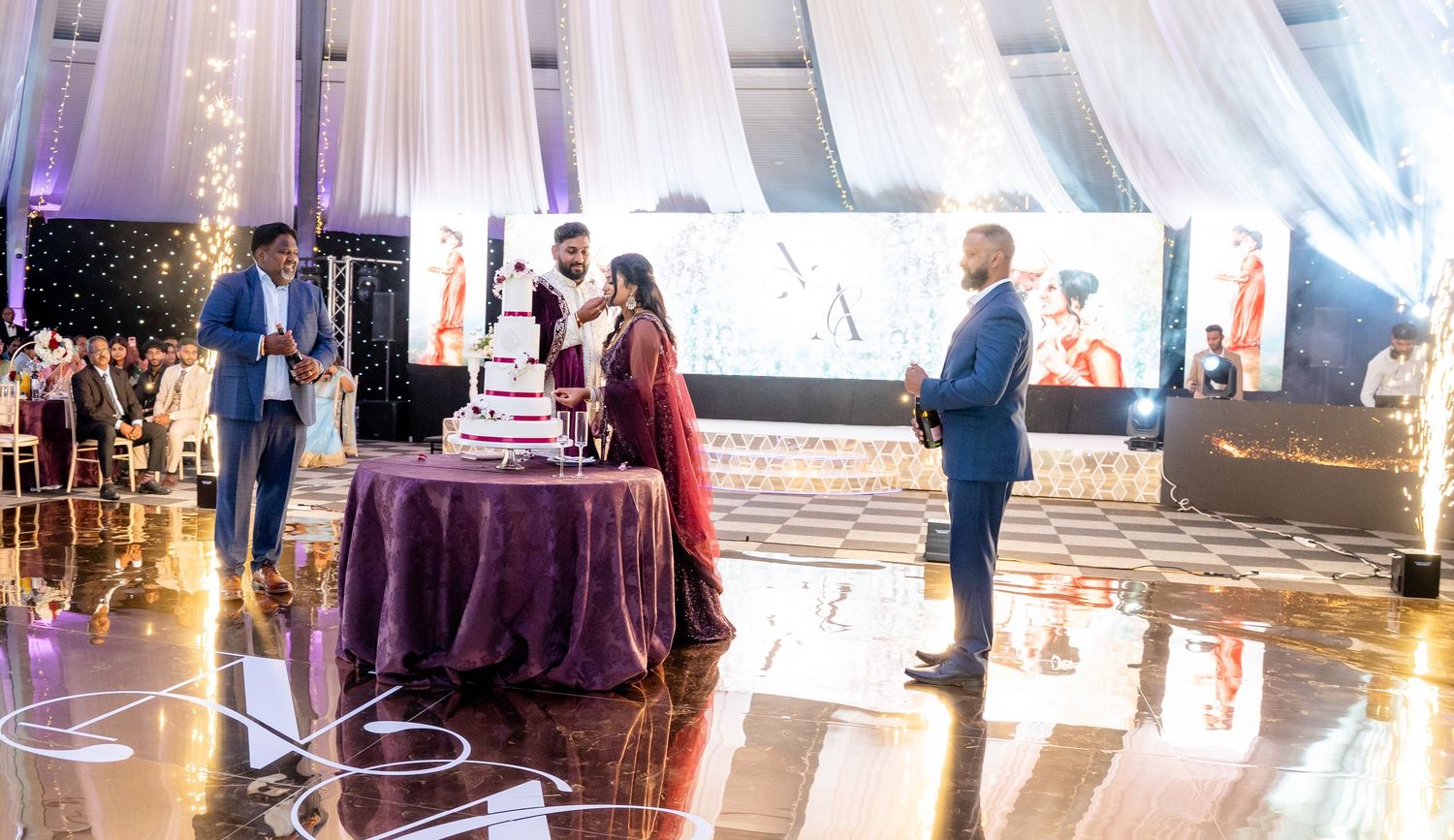 Indian bride and groom cutting tiered wedding cake on dance floor in beautifully decorated reception venue.