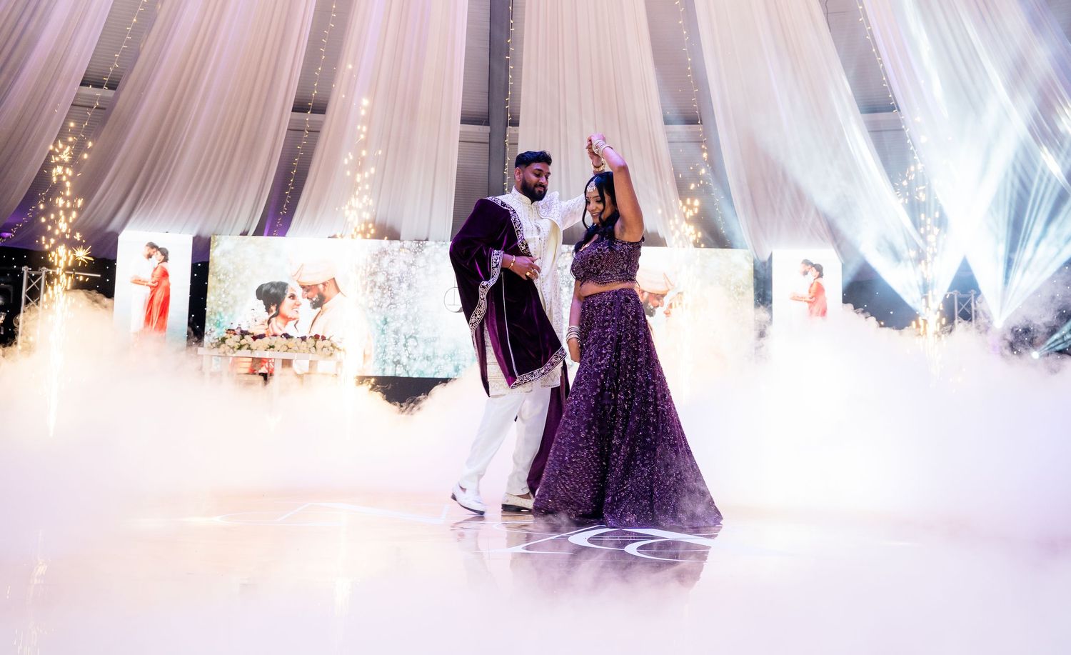Couple shares first dance in purple wedding attire on a white dance floor with dramatic fog and draped fabric backdrop.