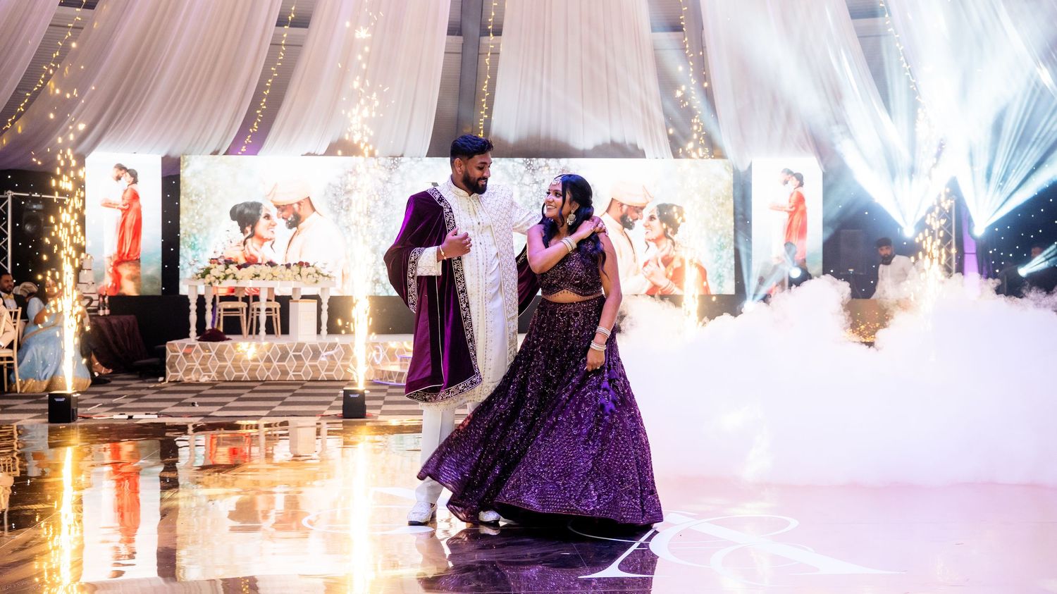 Couple shares first dance at Indian wedding reception with purple lehenga, sparklers, and elegant floral backdrop.