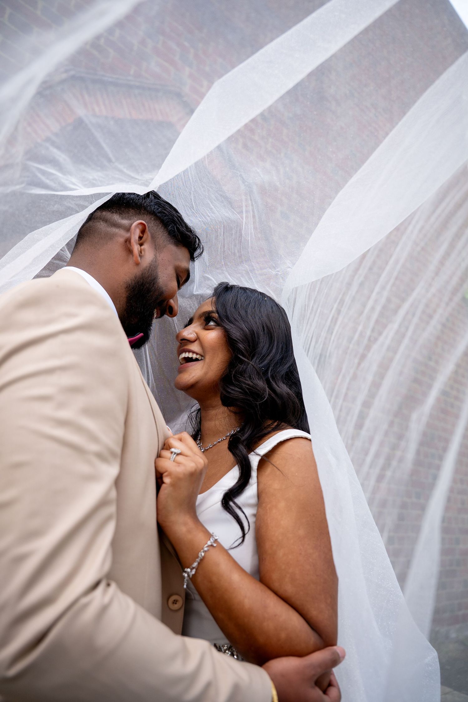 Bride smiles joyfully at groom beneath a billowing white veil in a romantic close-up wedding portrait.