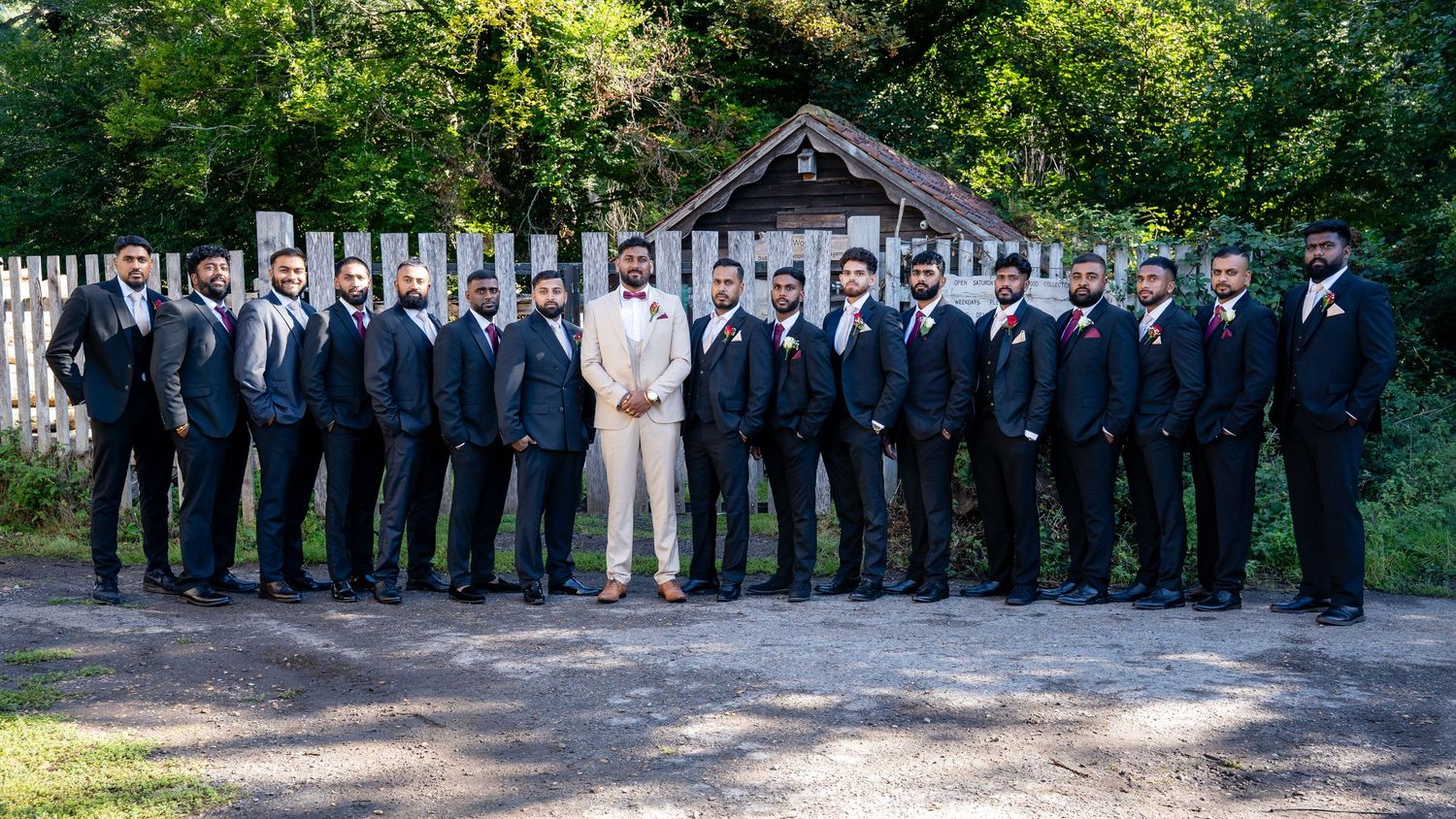 Groom in beige suit stands with large groomsmen party in black suits at outdoor wedding venue.