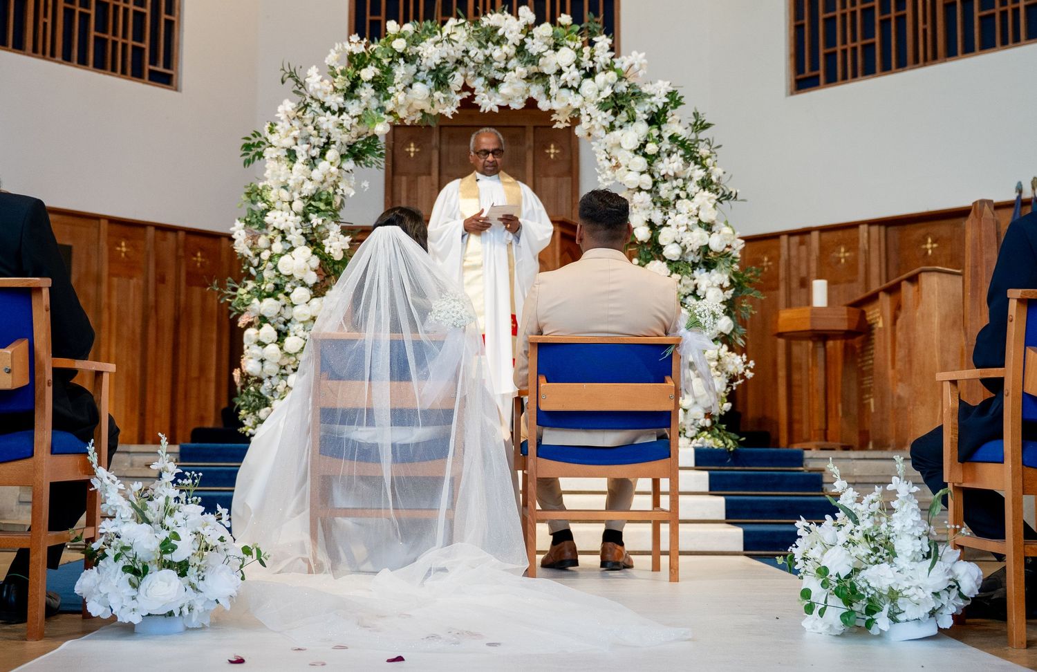Elegant church wedding ceremony setup with white floral arch, blue ribbon-decorated chairs, and flowing white fabric draping.