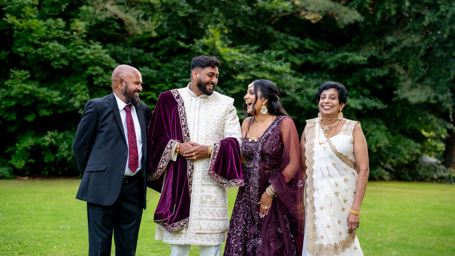 A groom in purple velvet sherwani poses with family members on a green lawn at a wedding.