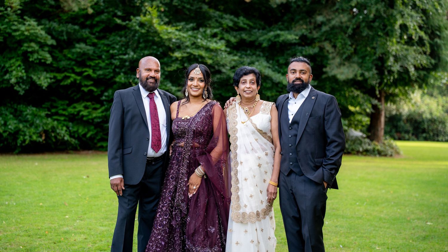 Four guests in elegant Indian wedding attire posing together on a lush green lawn at an outdoor celebration.