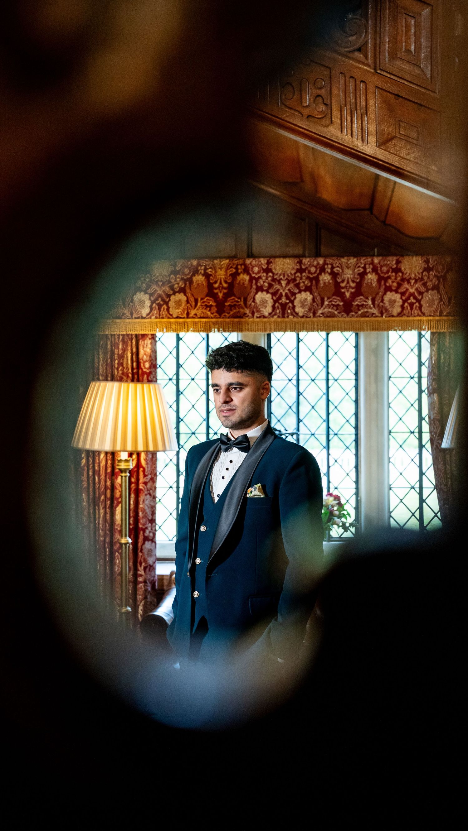 Groom in navy tuxedo with bow tie stands by leaded window in elegant wood-paneled wedding venue.