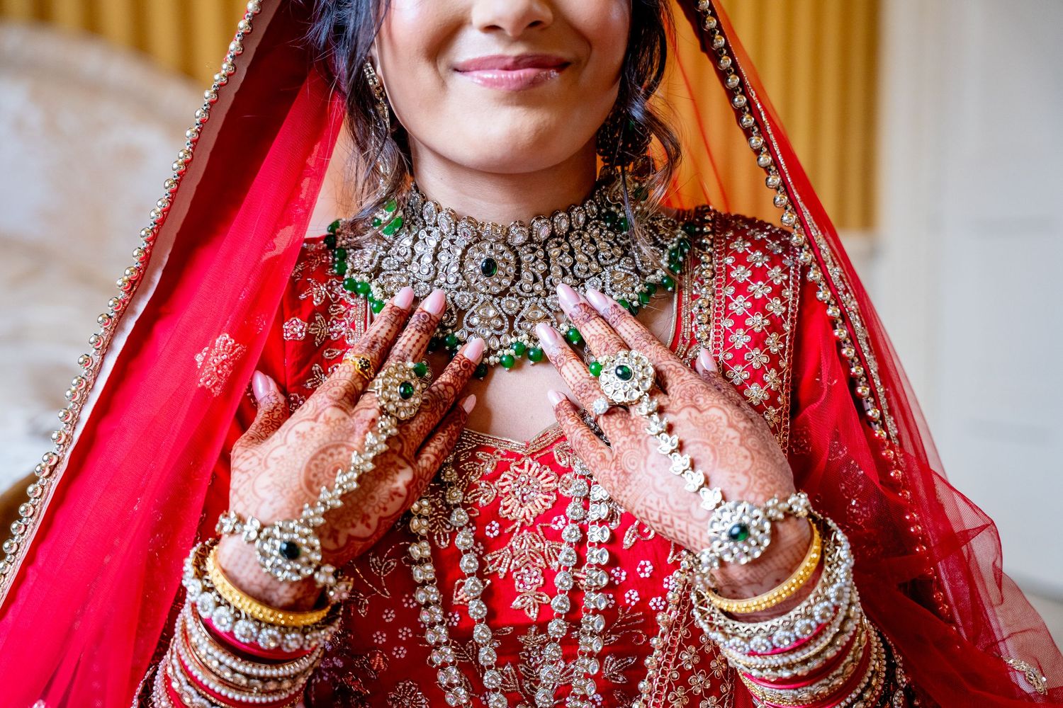 Indian bride in red lehenga showcasing elaborate necklace and mehndi-covered hands with green rings.
