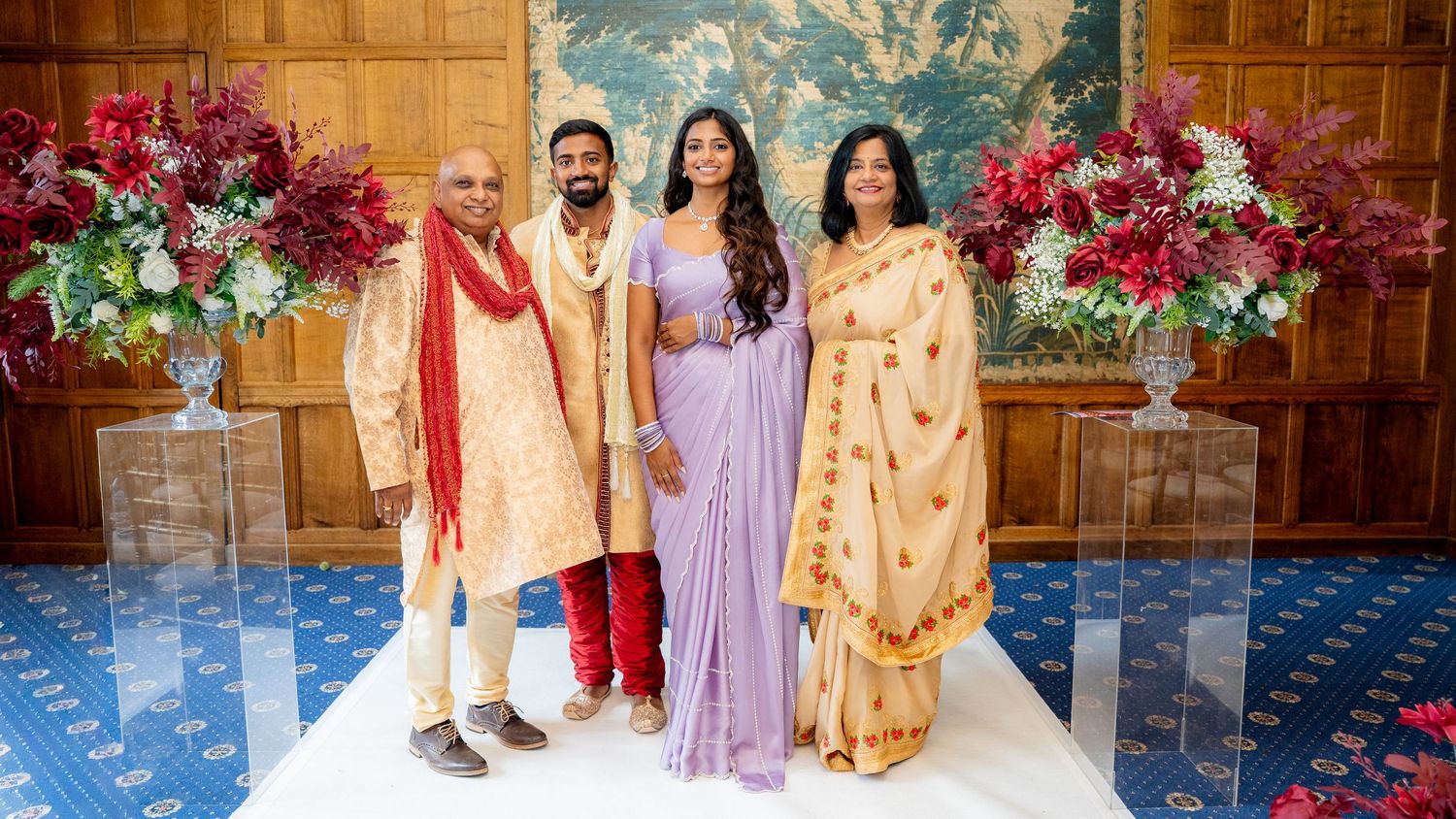 Four people in traditional Indian attire pose together at a wedding celebration with floral arrangements.