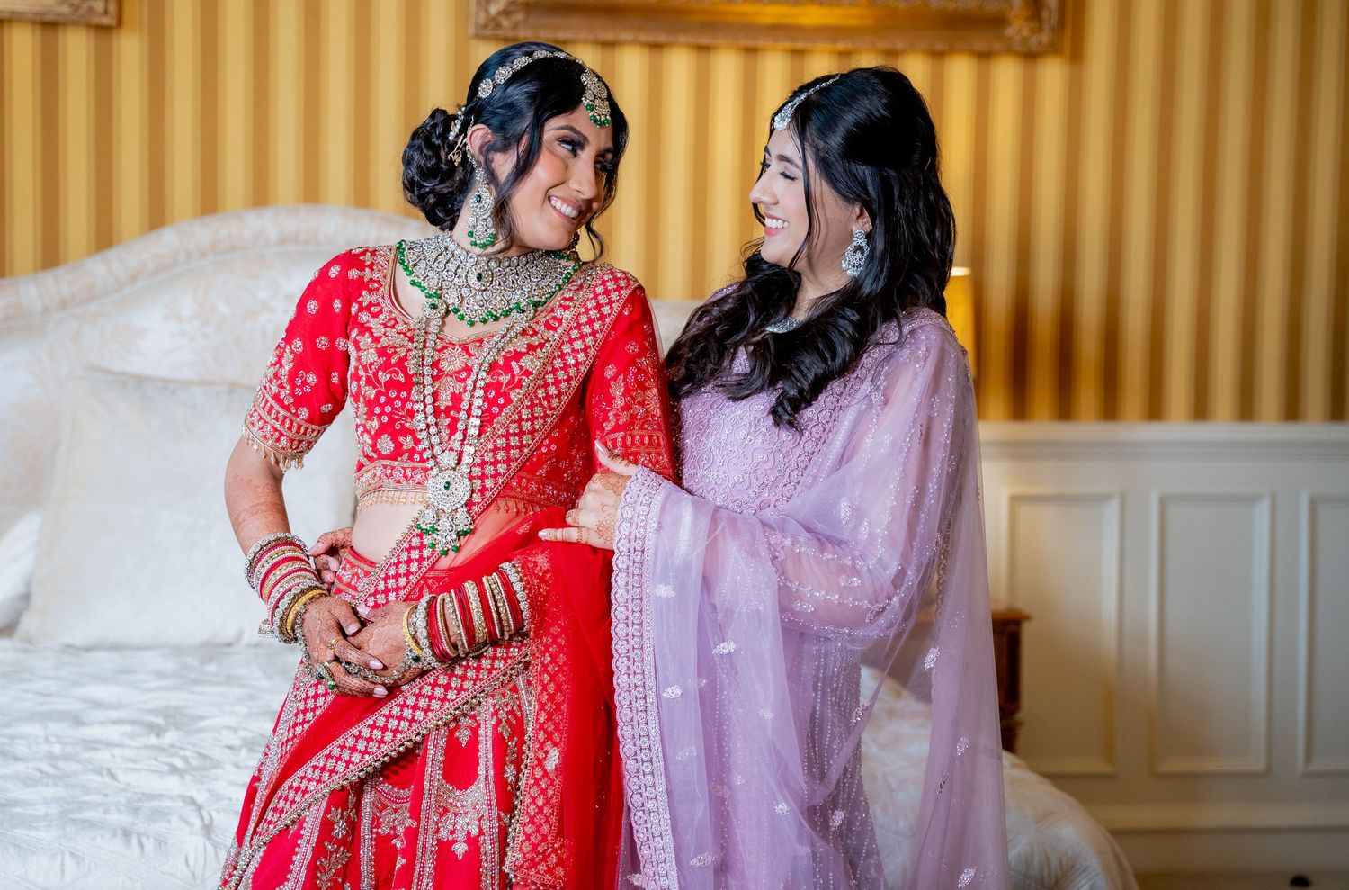 Indian bride in red lehenga laughs joyfully with bridesmaid in lavender outfit on white bed.