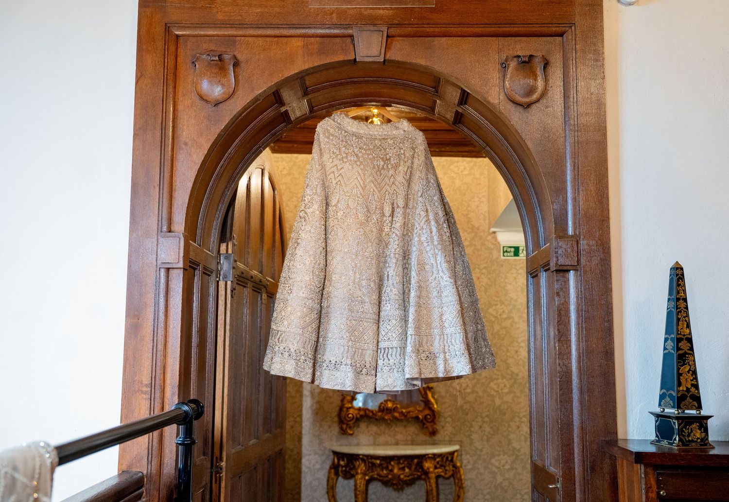 Ornate beaded silver dress hanging in an elegant wooden arched doorway with decorative molding in a historic interior.