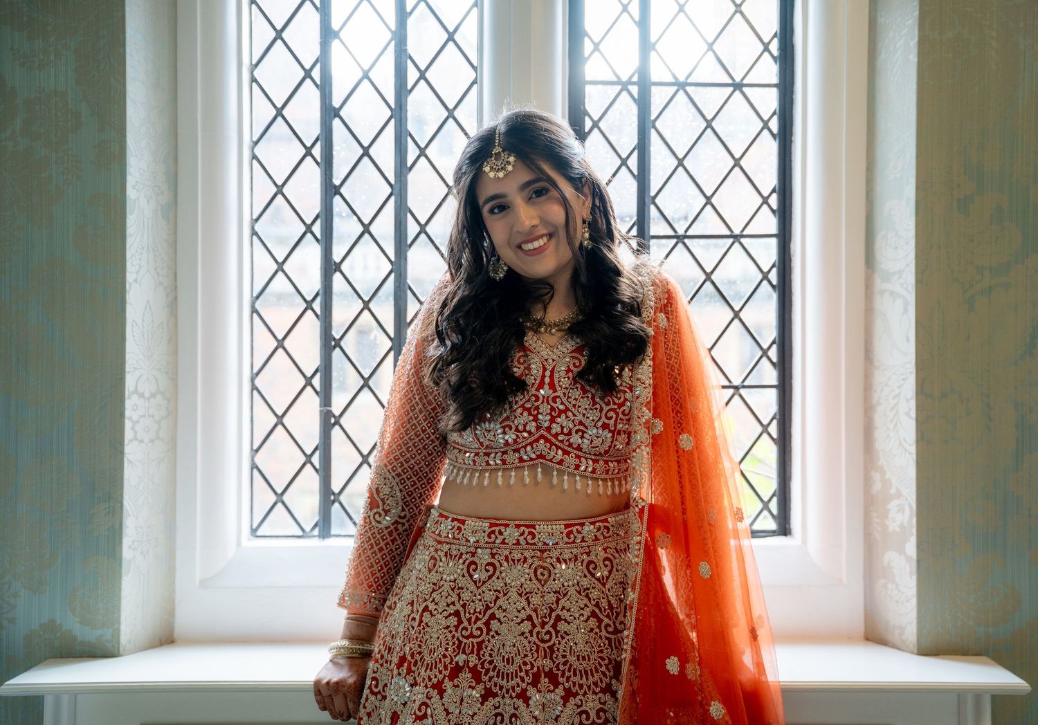 Smiling Indian bride in red and orange lehenga sits by diamond-paned window in bridal suite.