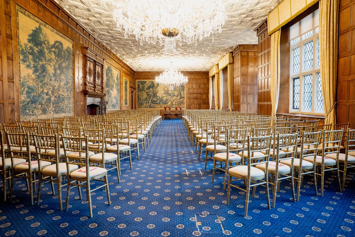Elegant ballroom with gold Chiavari chairs, blue carpet, crystal chandelier, and tapestry walls set for a formal ceremony.