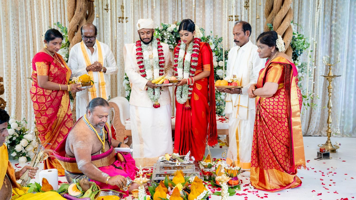 Hindu wedding ceremony with priest conducting rituals as family members hold lamps blessing the couple on mandap.