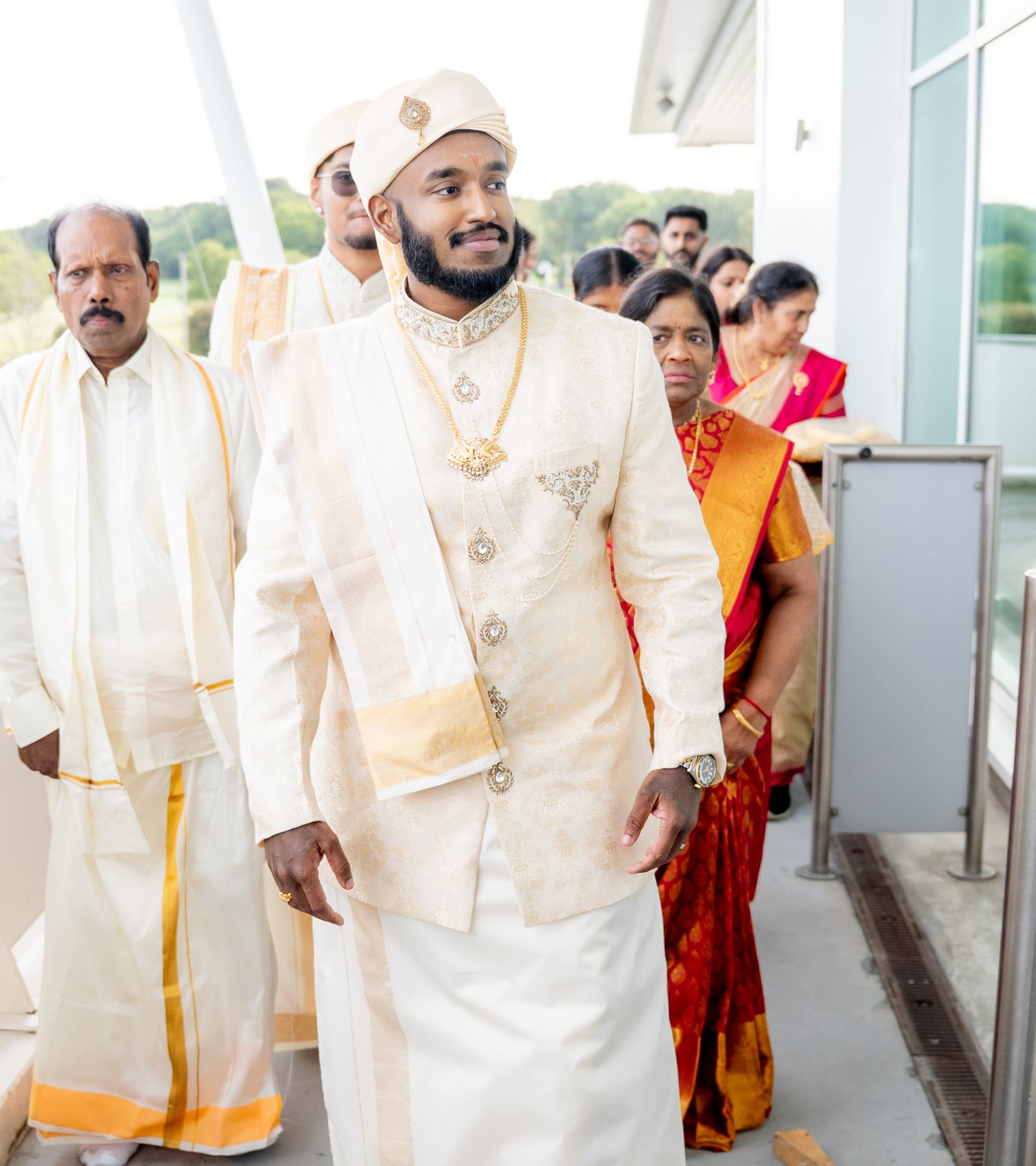 Groom in ornate white and gold Indian wedding sherwani with turban walks through a modern wedding venue.