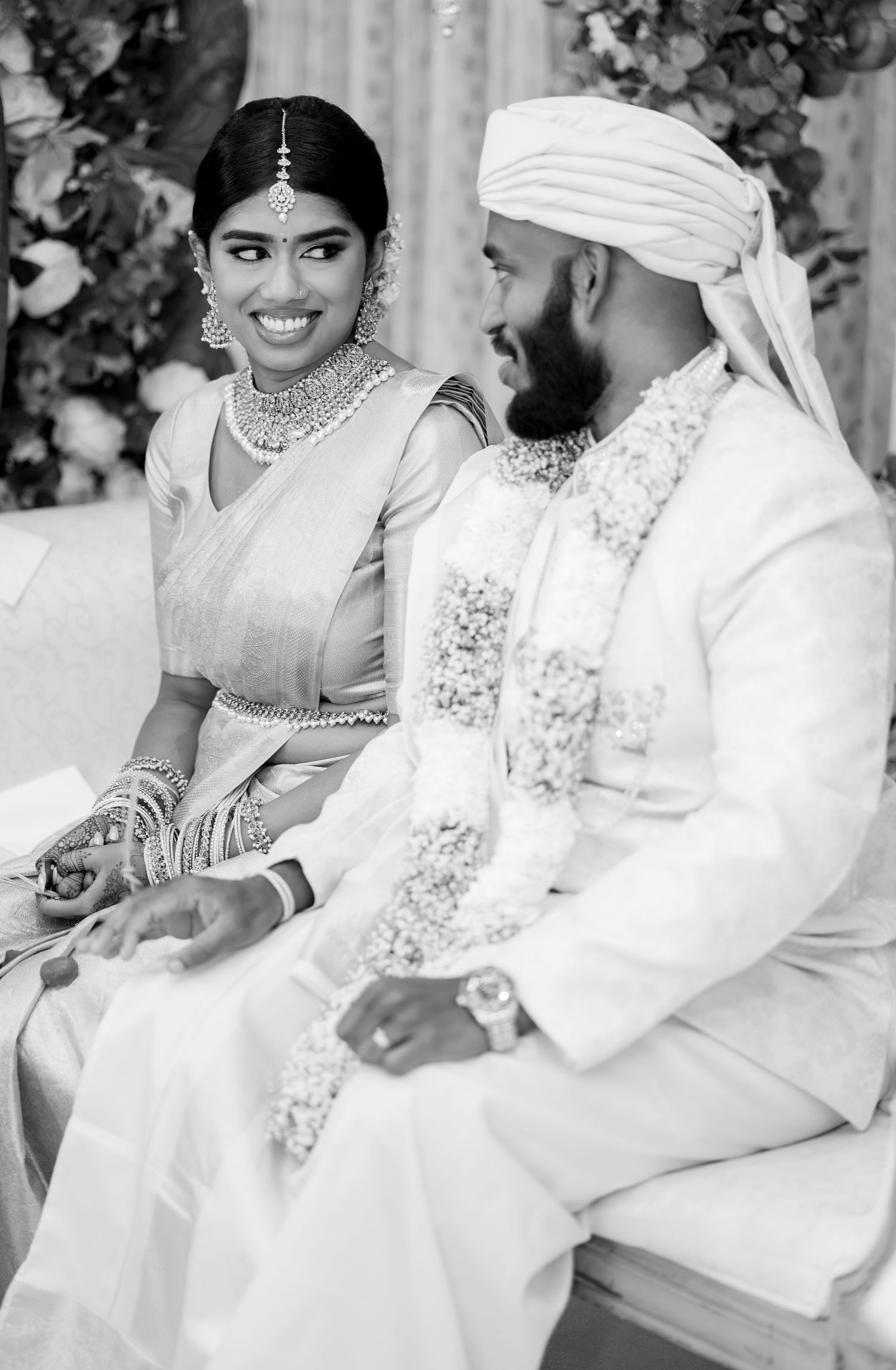 Black and white portrait of South Indian bride smiling at groom during traditional wedding ceremony at floral mandap.