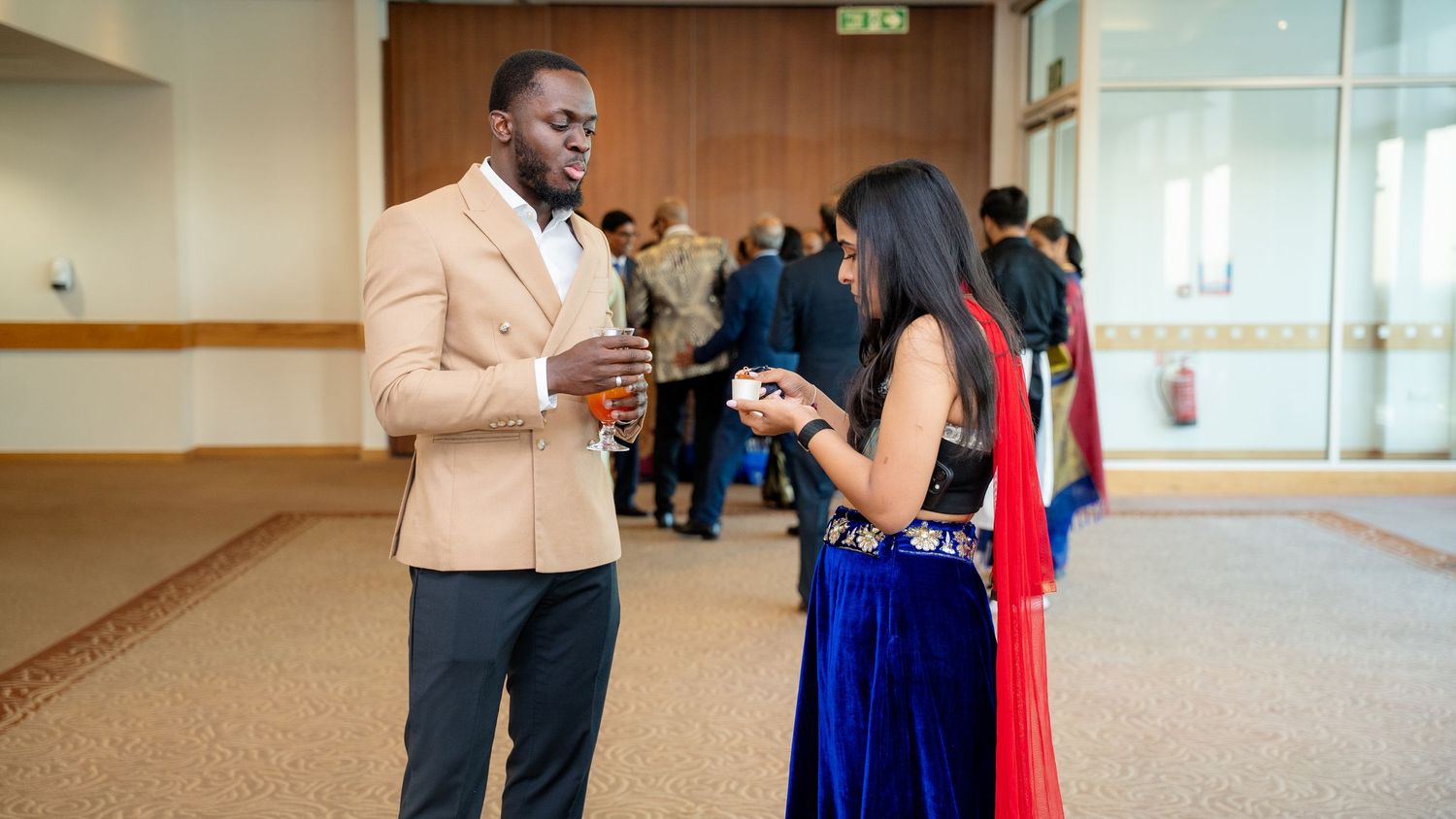 Man in beige blazer and woman in blue saree conversing at a formal gathering event.