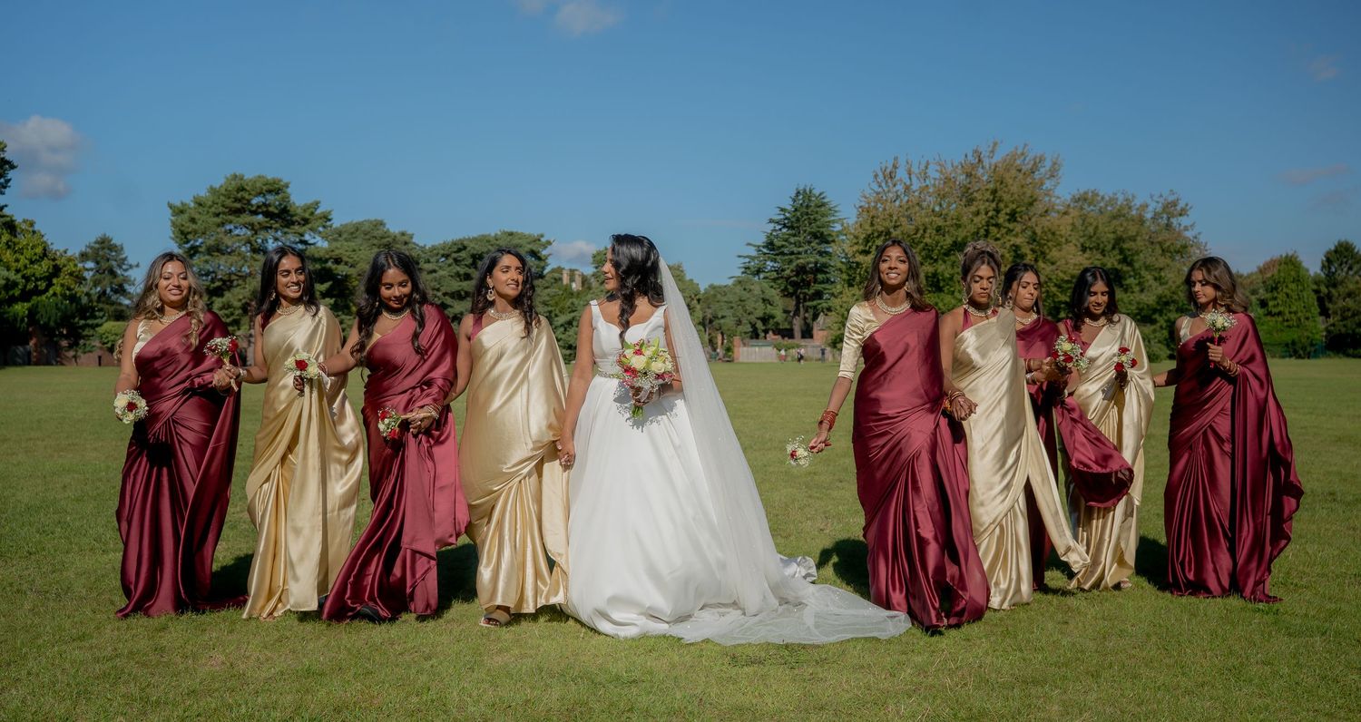 Bride and bridesmaids in burgundy and gold dresses posing outdoors on green lawn under blue sky.