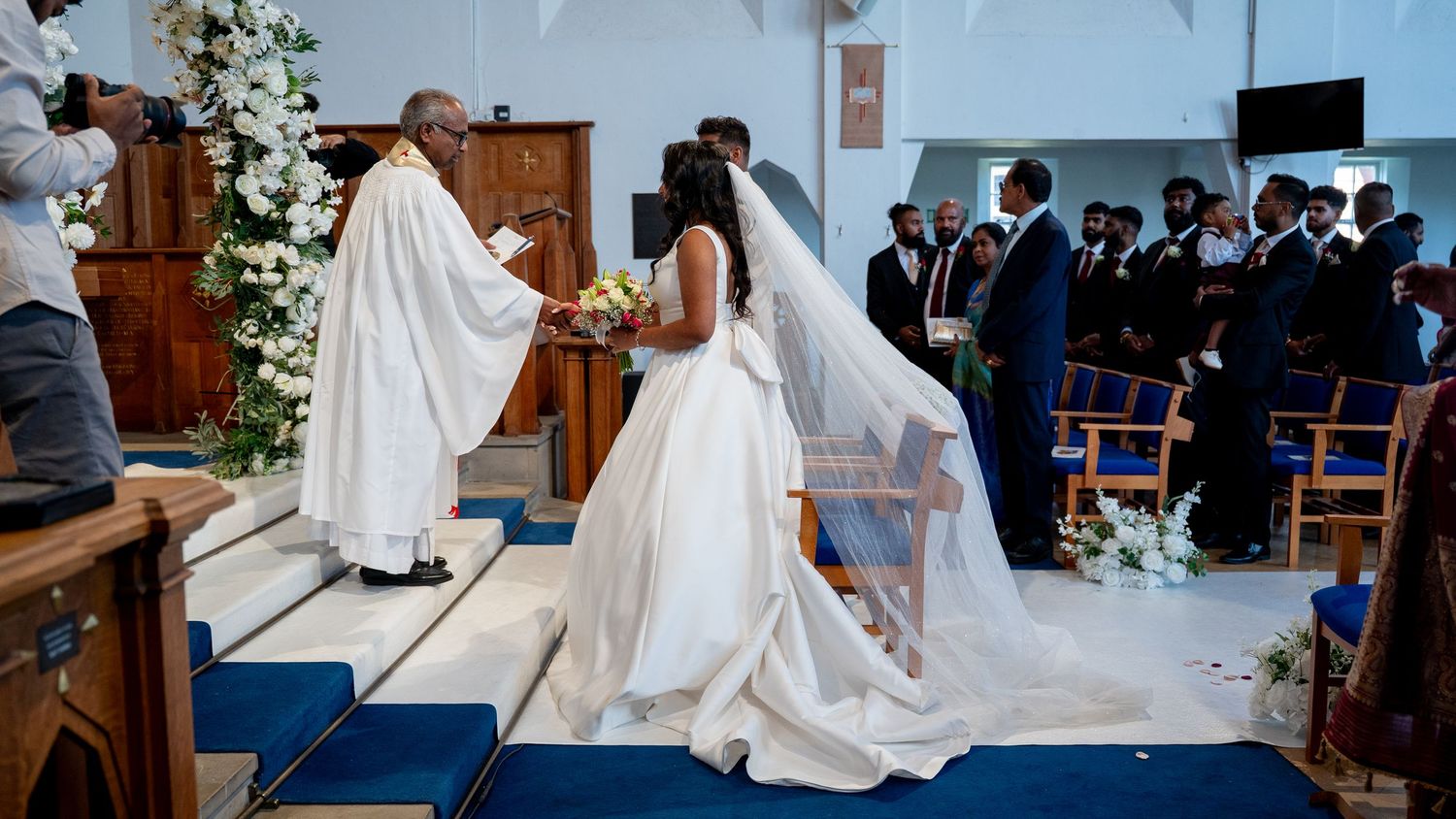 Bride in white gown and veil approaches priest at flower-decorated church altar during ceremony.