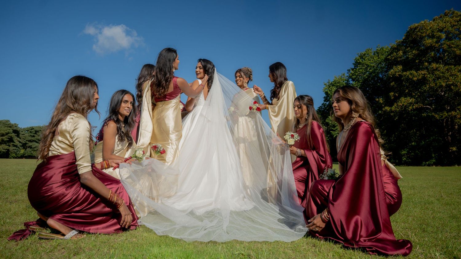Bride in white wedding gown surrounded by bridesmaids in burgundy dresses on sunny lawn with trees in background.