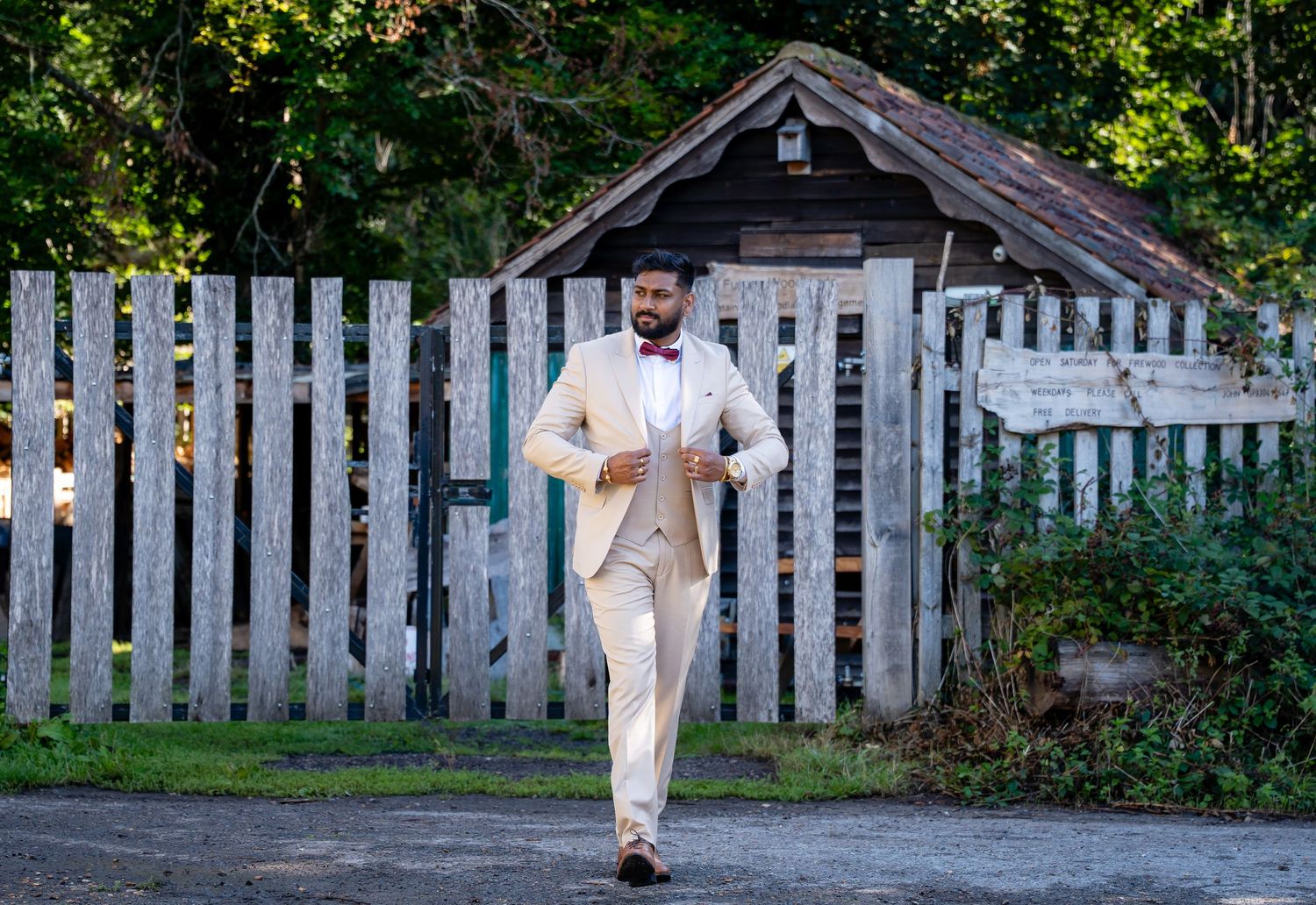 Groom in cream suit walking confidently down a path near a wooden fence and shed.