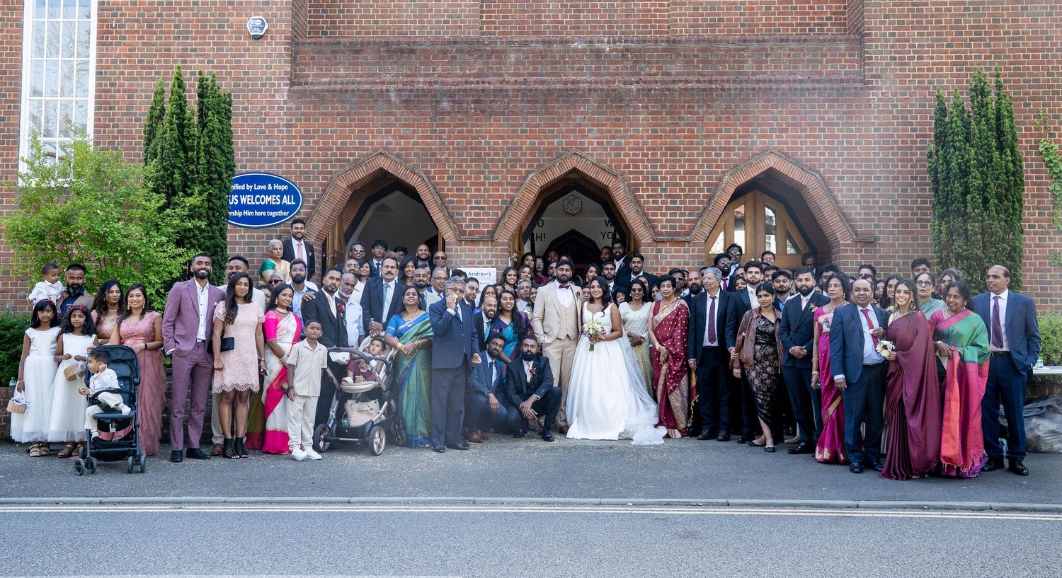 Large wedding group photo outside a brick church, bride and groom surrounded by guests in colorful formal attire.