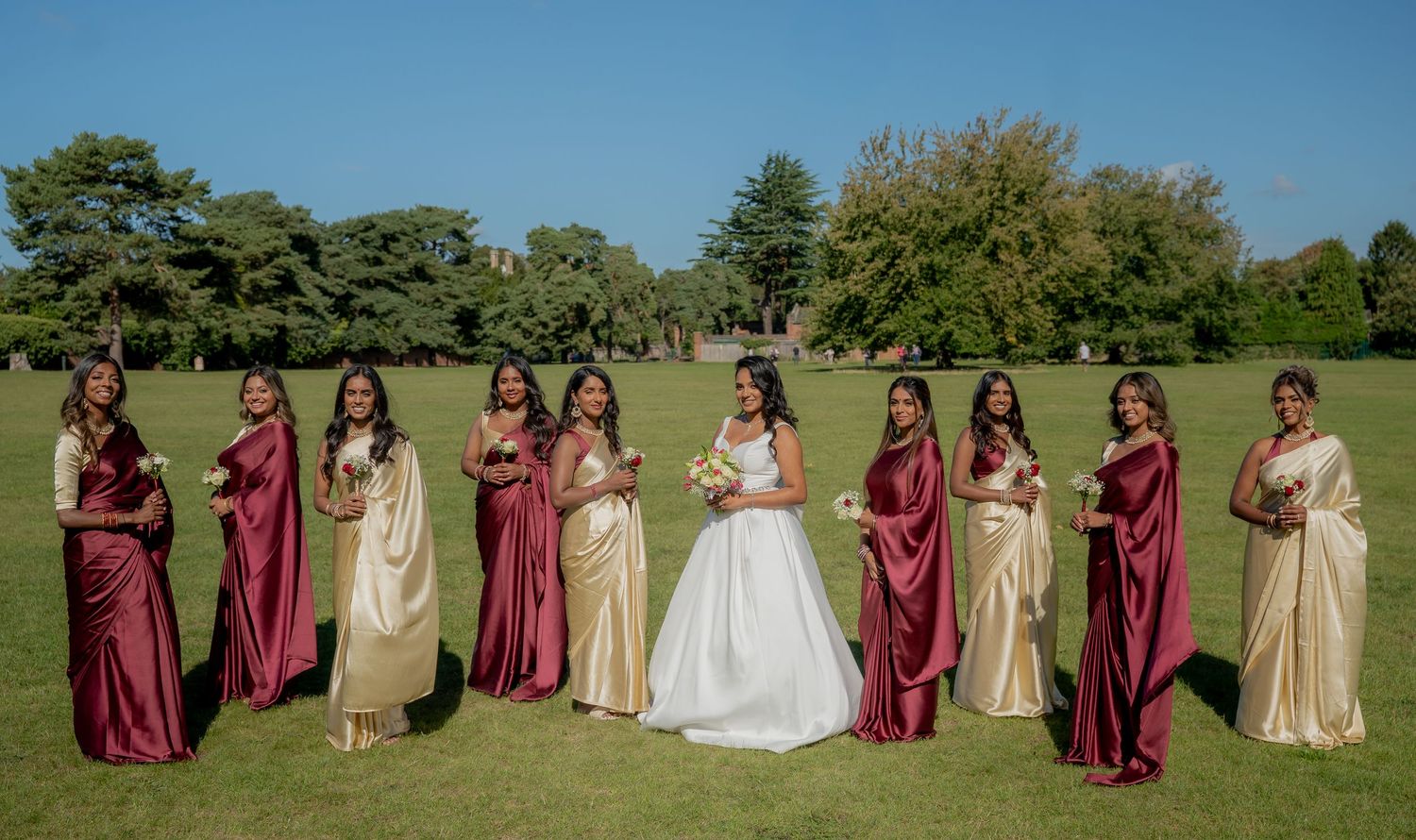 Bride in white gown poses with bridesmaids in burgundy and gold saris on a sunny green lawn.