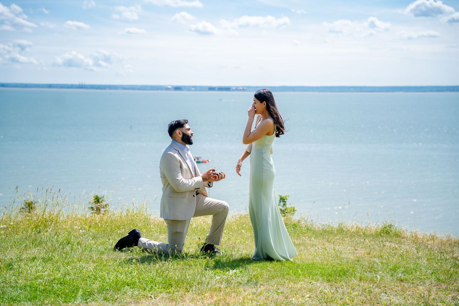 Man proposing on one knee to woman in white dress with ocean backdrop on sunny day.