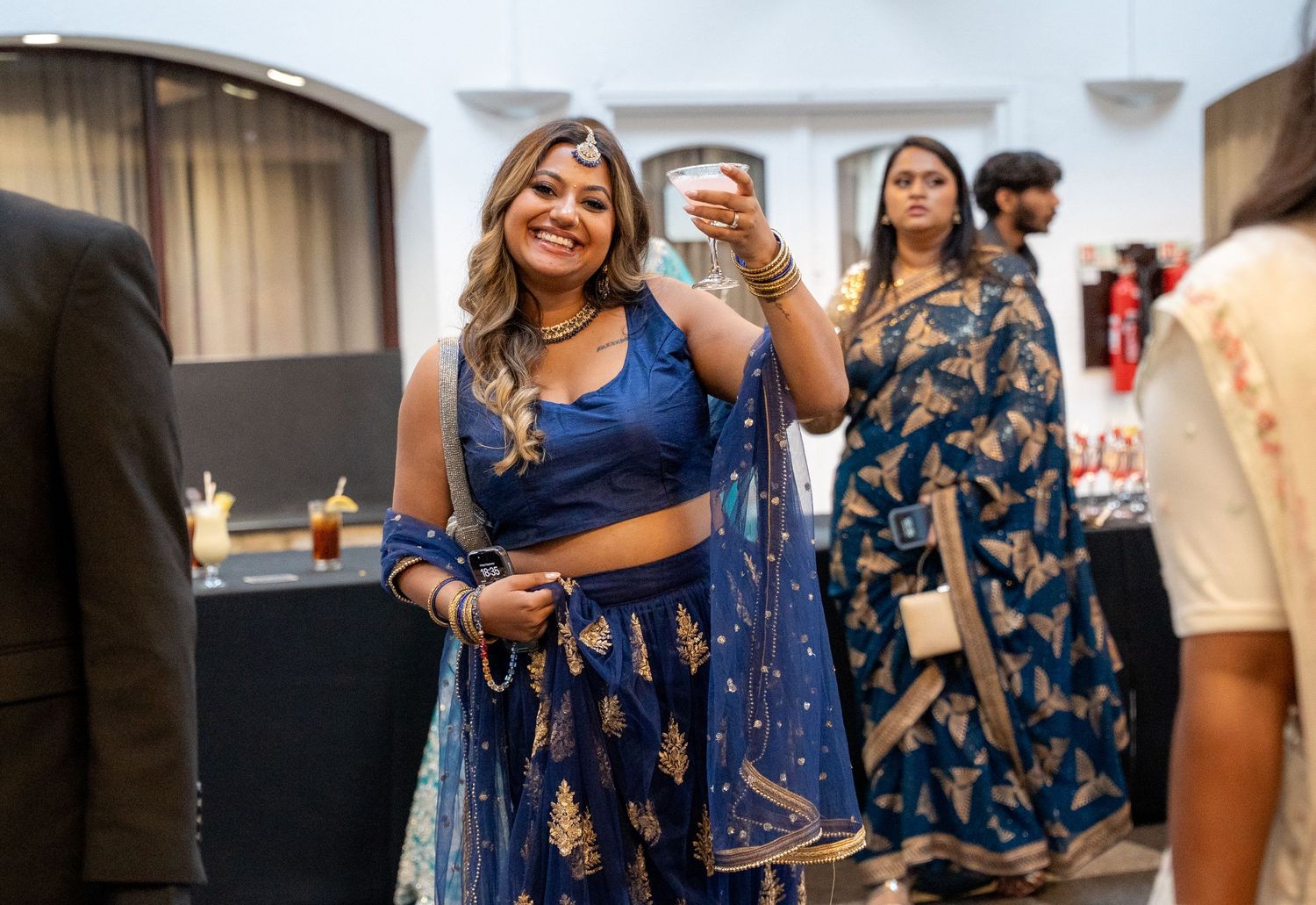 A woman in a blue embroidered lehenga smiles and raises a glass at an Indian wedding celebration.