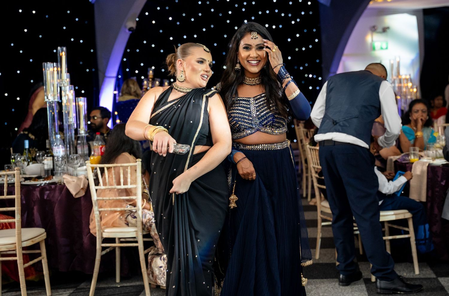 Two women in elegant Indian attire smile and pose together at a formal banquet event with decorative lighting.