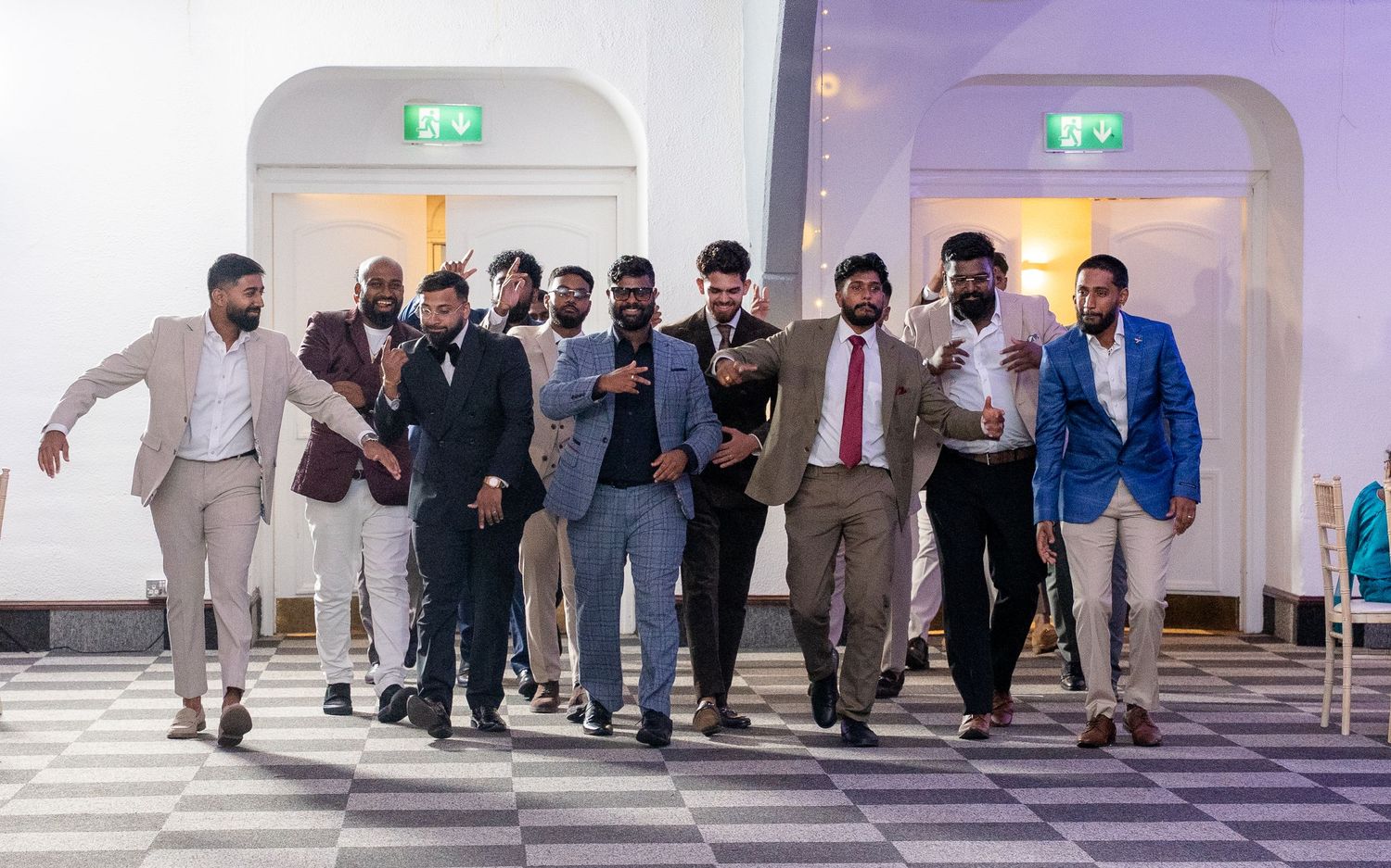 Group of groomsmen in suits walking confidently together inside elegant wedding venue reception hall.