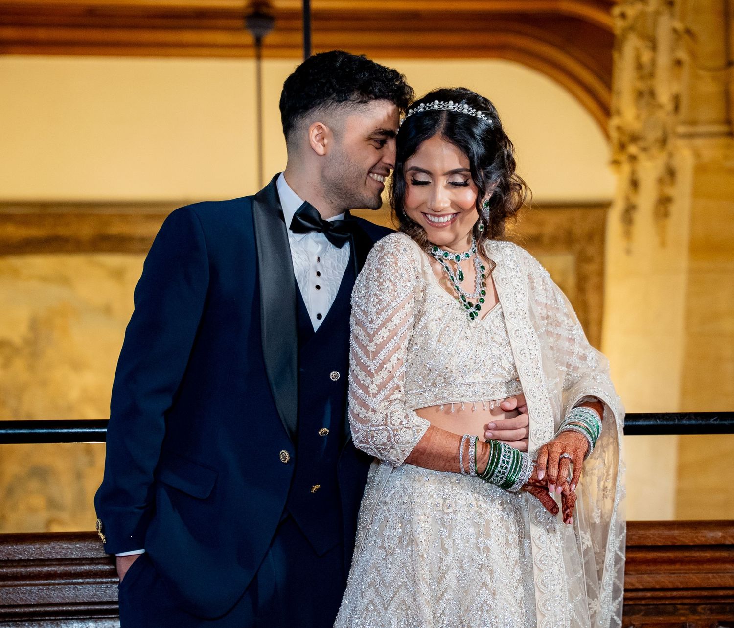 Couple in elegant wedding attire, groom in navy tuxedo, bride in white embroidered lehenga with green jewelry, posing indoors.
