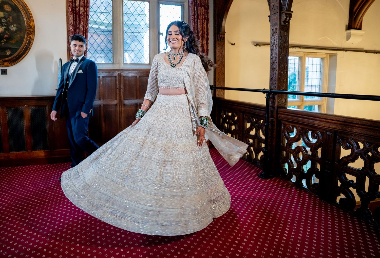 Bride twirls white embroidered lehenga on grand staircase as groom watches in historic wedding venue.