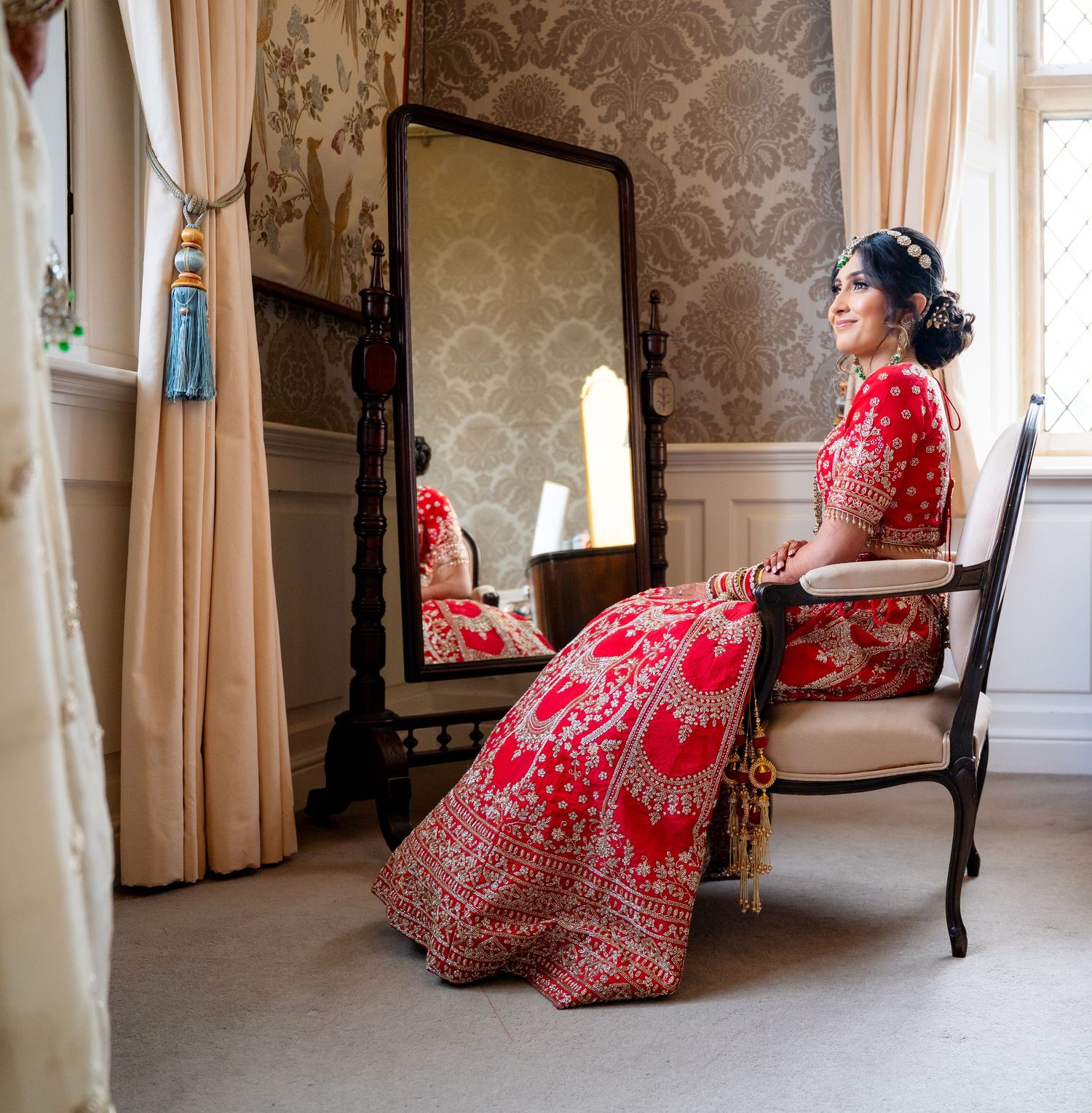 Indian bride in red lehenga seated beside full-length mirror in elegant wallpapered dressing room.