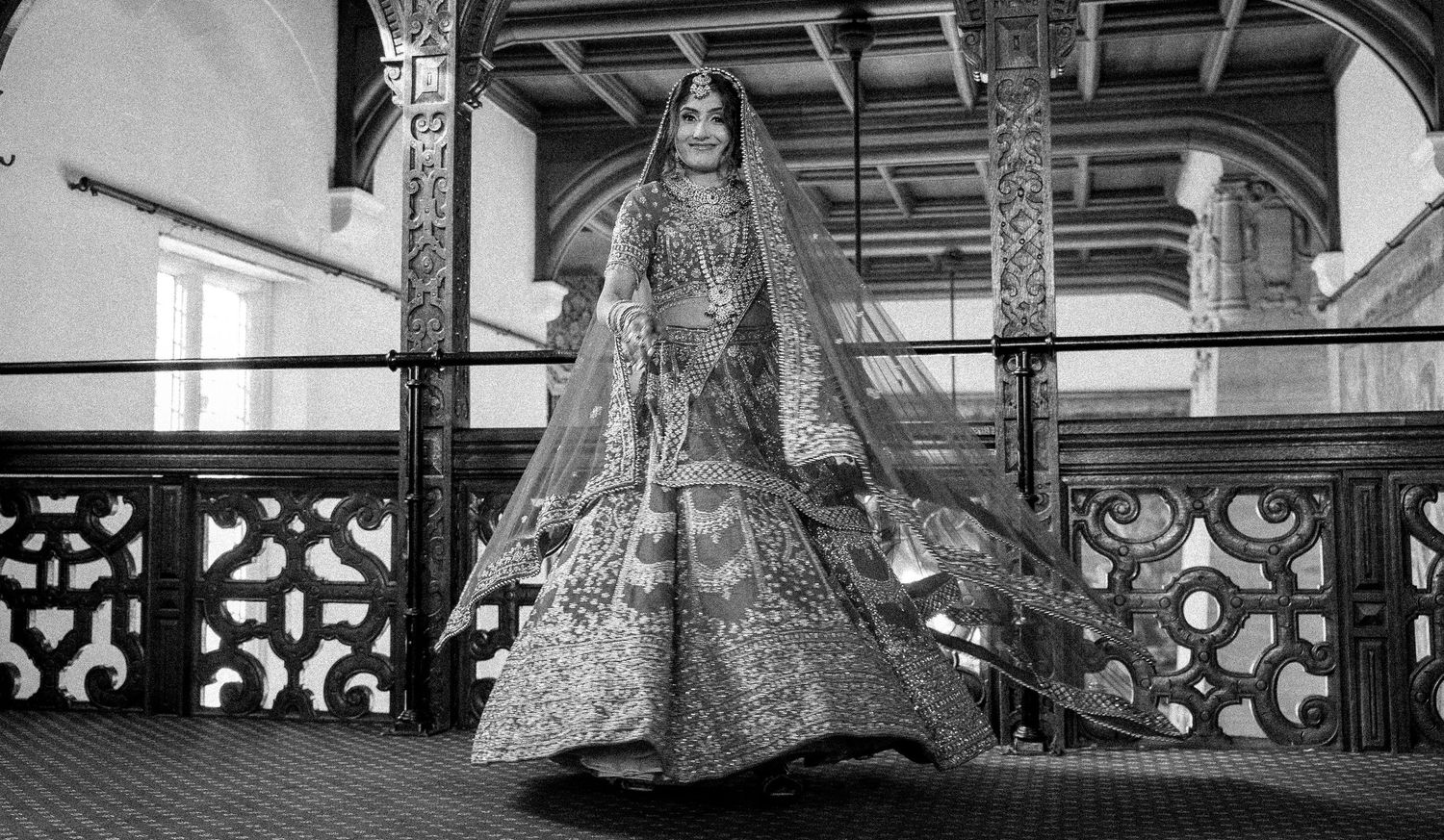 Black and white photo of Indian bride twirling in embroidered lehenga with veil on ornate balcony.