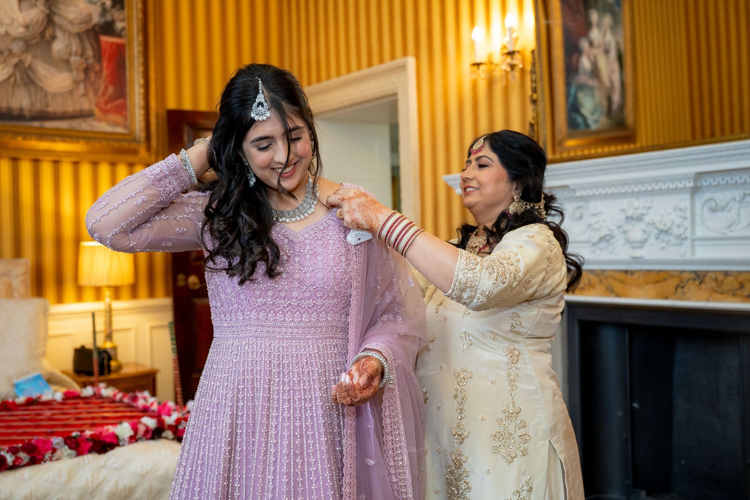 Two women in elegant South Asian attire help each other prepare, one in lavender anarkali and the other in ivory suit, in a grand room.