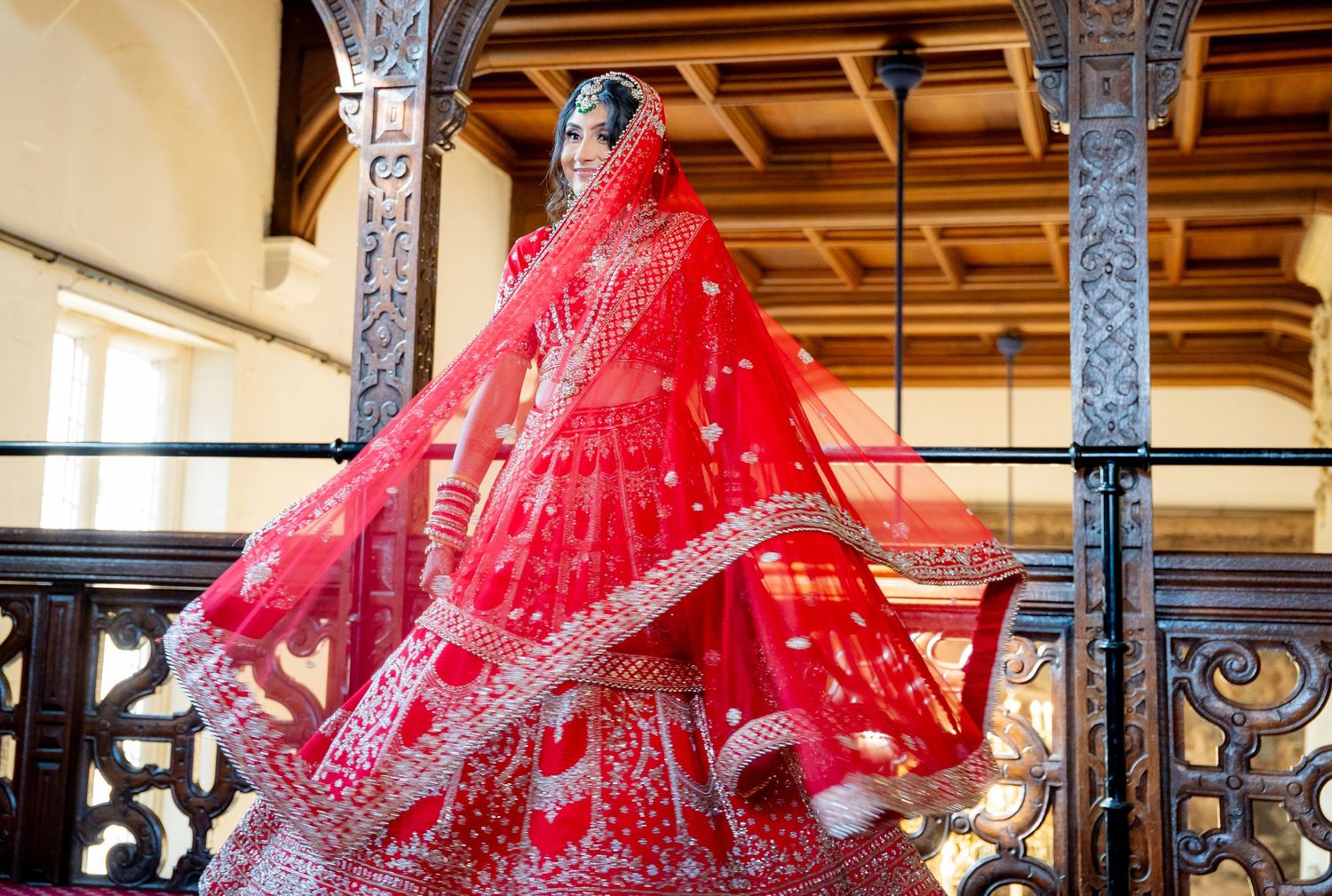 Indian bride twirling in a stunning red lehenga with embroidered dupatta on ornate balcony.