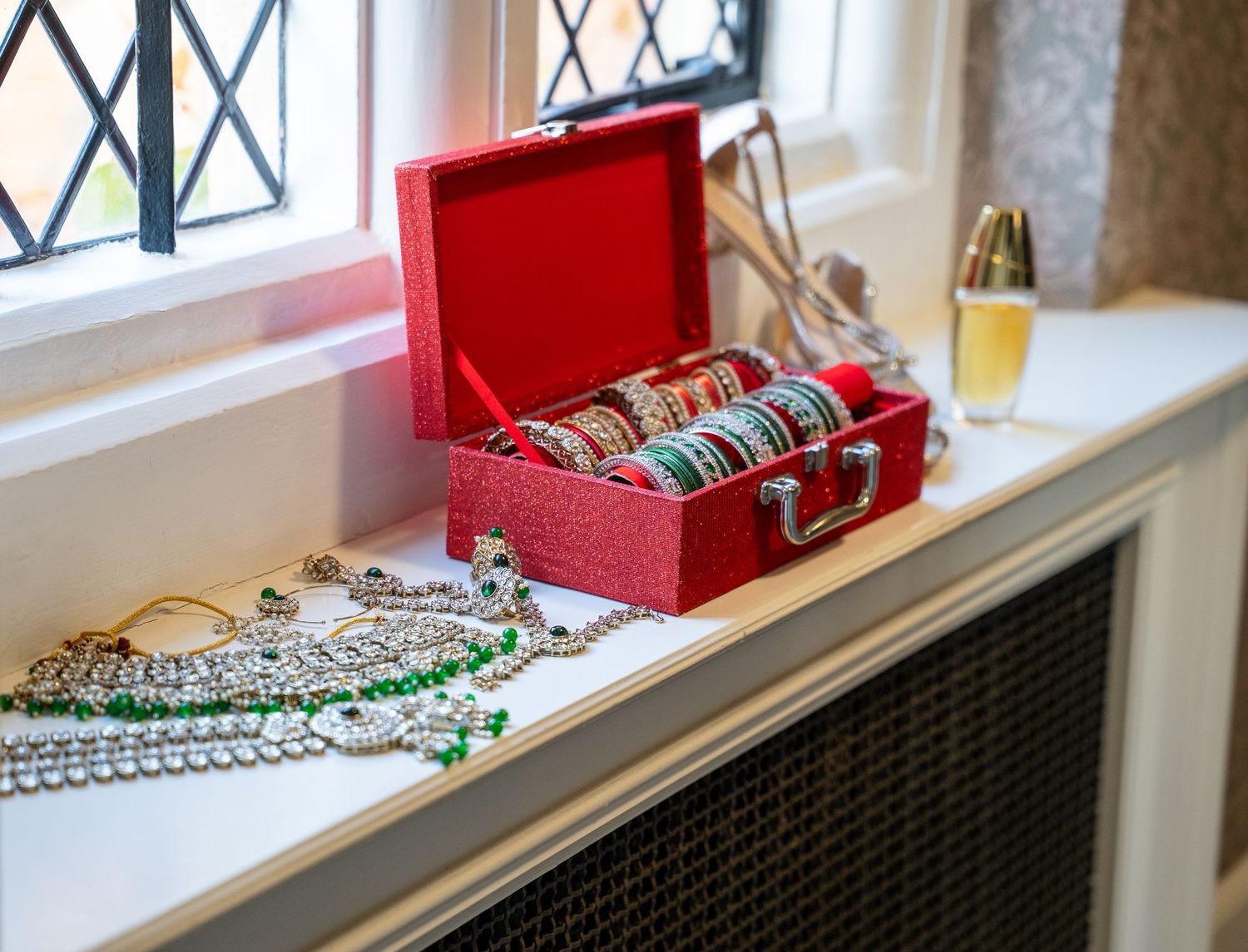 Red velvet jewelry box with bangles, pearl necklaces, and emerald beaded chains displayed on a windowsill.